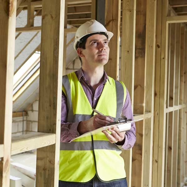 Construction worker in hard hat and safety vest inspecting framing, taking notes.