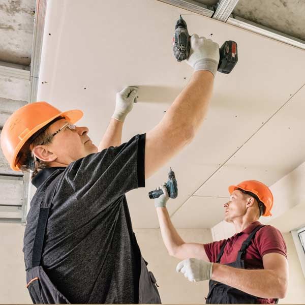 Two construction workers are installing drywall on a ceiling, one holding a drill, wearing safety gear.