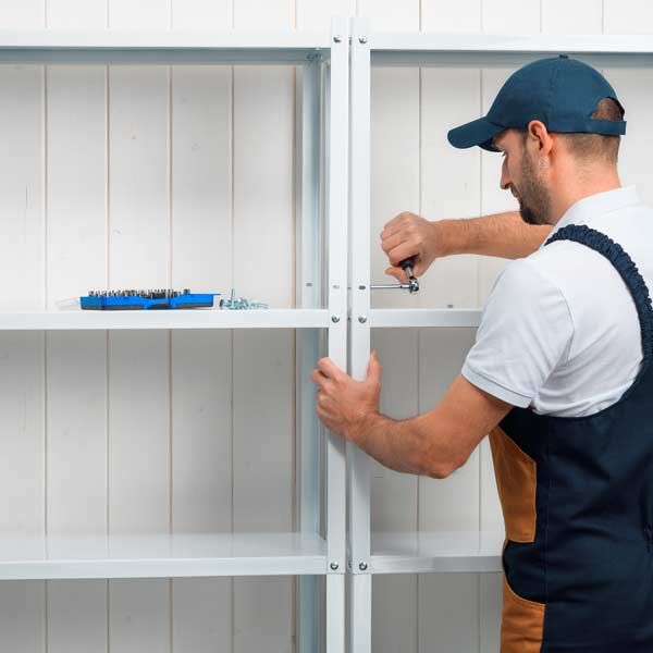 A man in a blue cap is assembling a white metal shelf unit with a screwdriver against a white wall. Shelving and Fixtures
