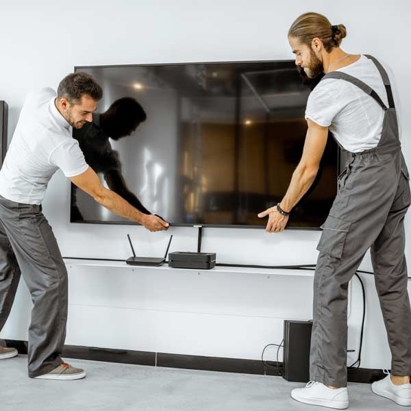Two men installing a large, flat-screen TV on a white wall. They're wearing work clothes and look focused TV Mounting Installation