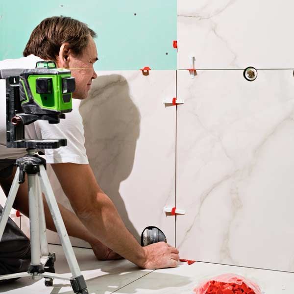 A handyman using a laser level to install white tiles on a wall and floor in a room. Professional Tile Installation and Repairs in Oceanside, Serving all of Nassau and Suffolk County, Long Island, for Over Twenty Years
