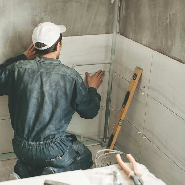 A worker in blue overalls installs tiles in a bathroom, using a level after clients Contacst Honest Hammer Handyman and Get Your Free Tiling Repair or Installation Consultation Today