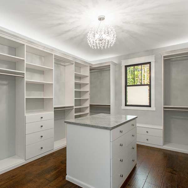 White walk-in closet with built-in shelves, drawers, and an island. Dark wood floor, a small window, and a chandelier.