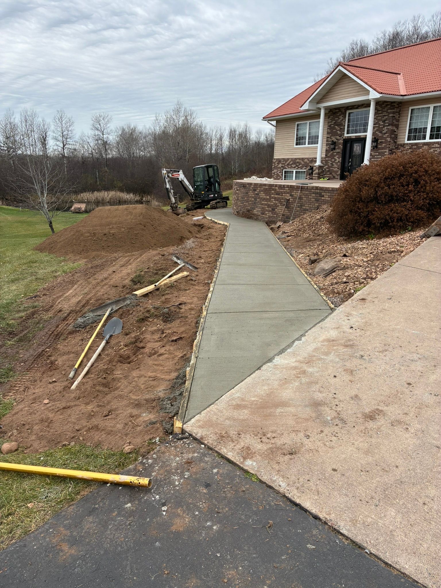 A newly poured concrete walkway connects to a driveway in front of a stone house, with a small excavator in the background.