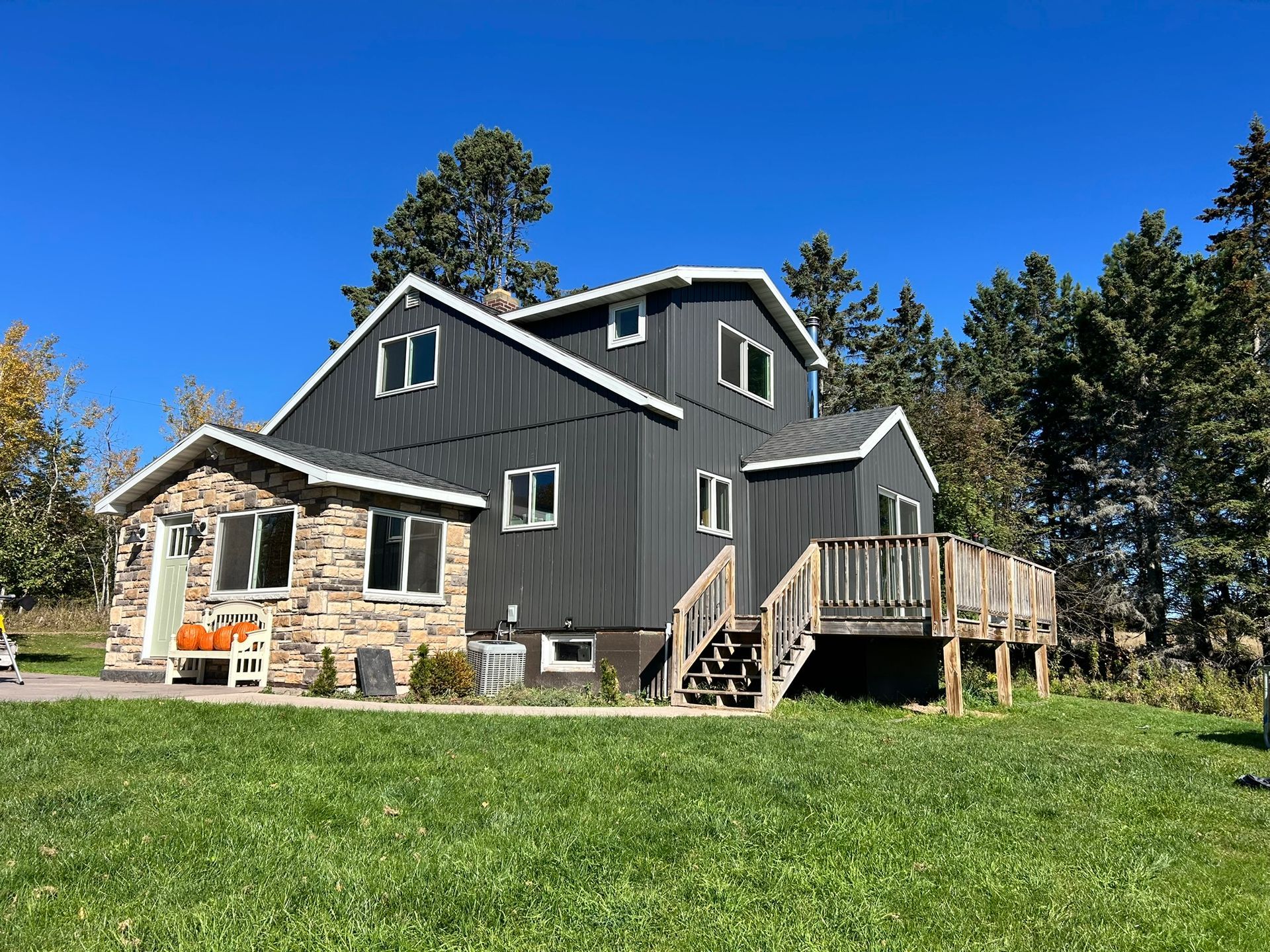 A two-story dark gray home with vertical siding and a stone-clad side porch under a clear blue sky.