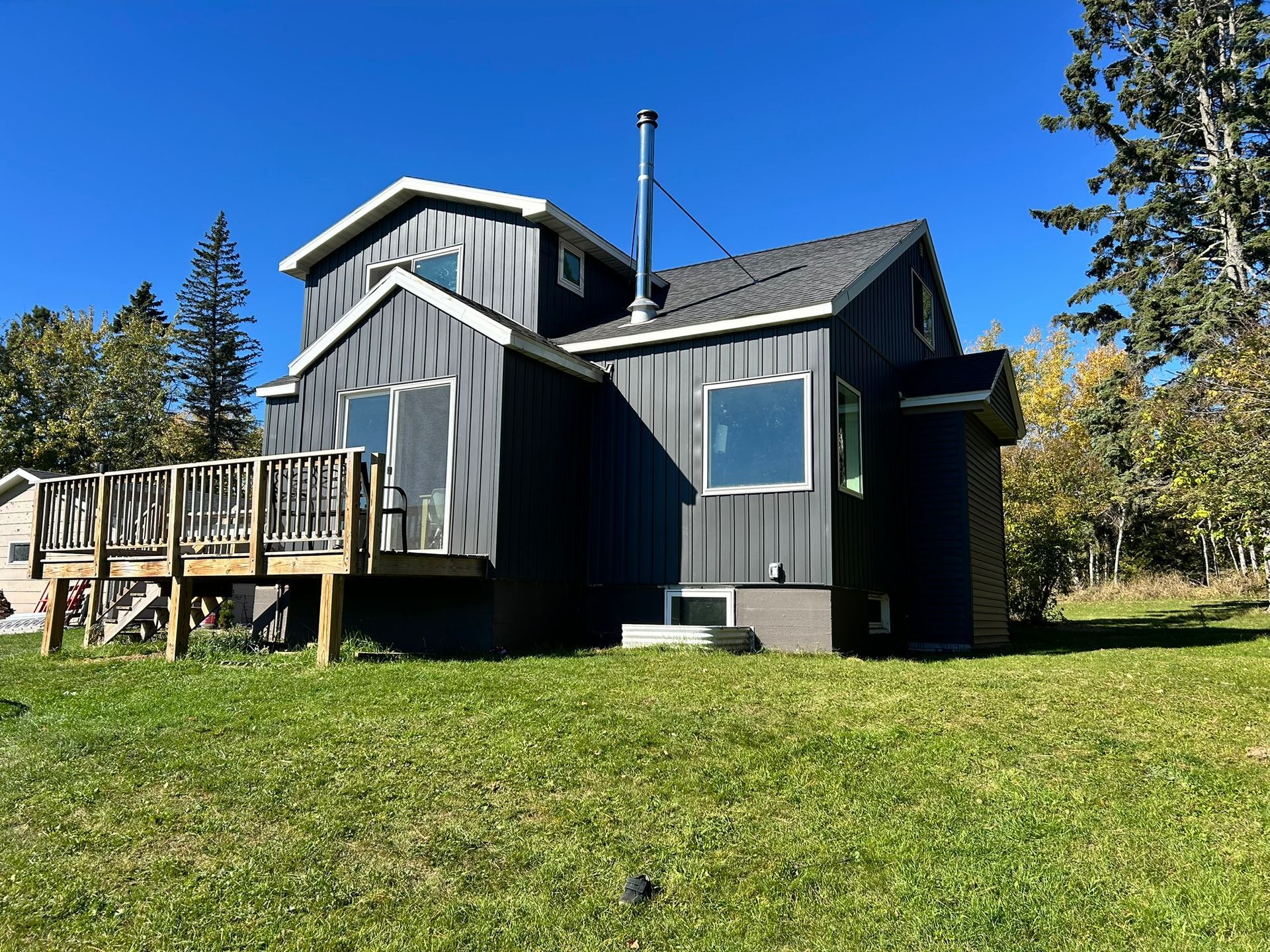 A dark gray house with a wooden deck and metal siding stands in a green grassy yard under a clear blue sky.