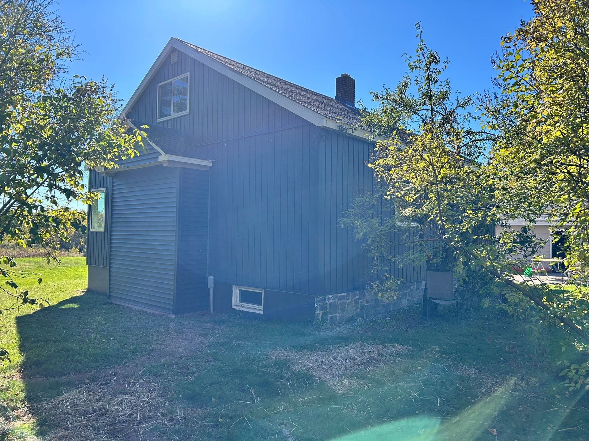 A dark-sided, two-story house with a small shed attached, surrounded by trees on a sunny day.