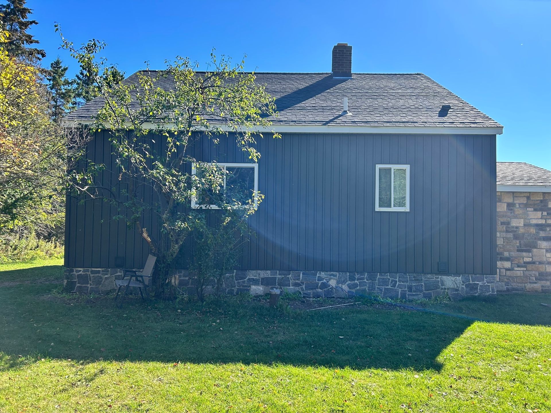A dark wooden house with a stone foundation, a chimney, and a small tree in front under a clear blue sky.