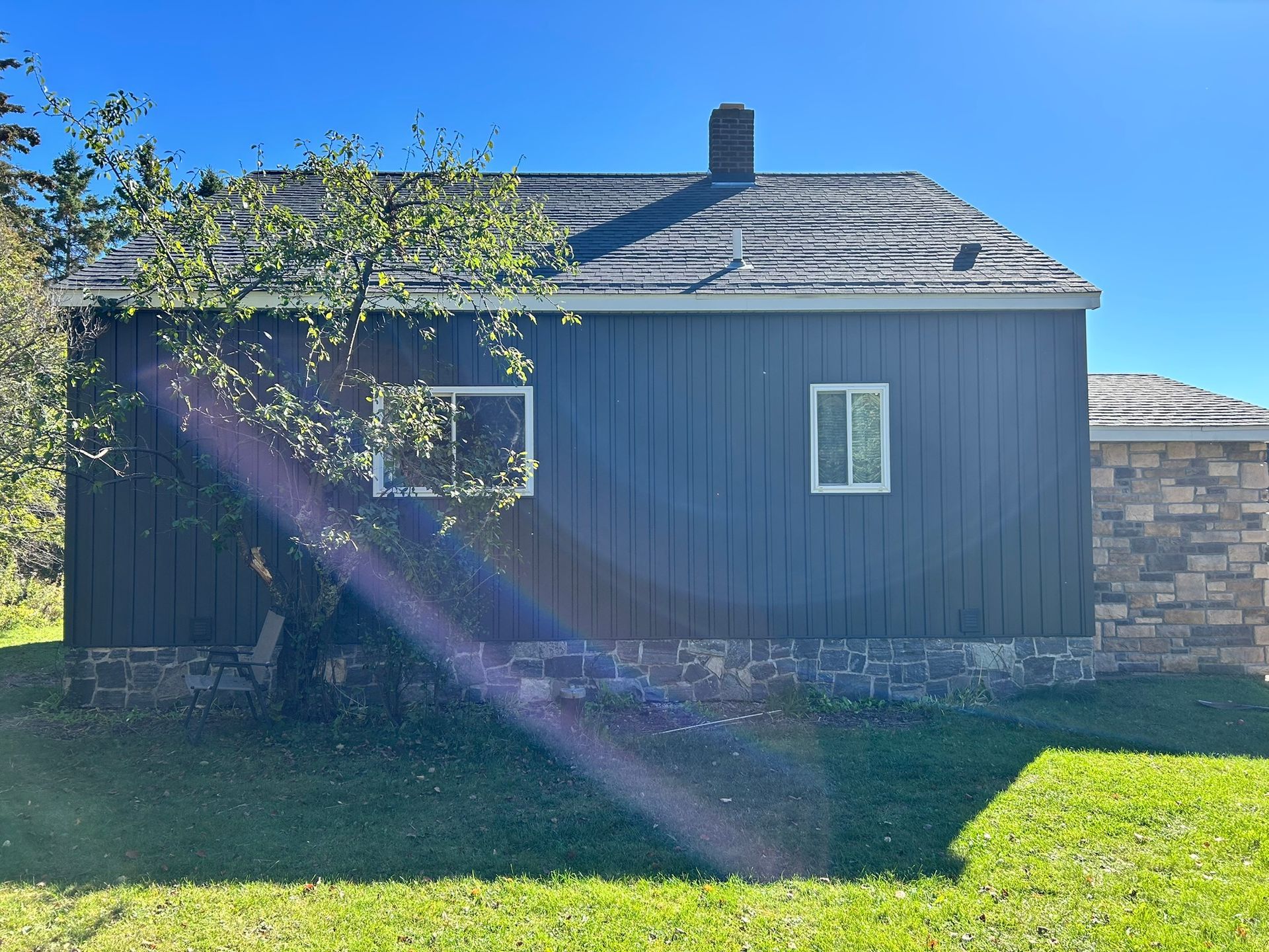 A dark vertical-sided cabin with a stone foundation, a dark shingled roof, and a stone extension, under a clear blue sky.