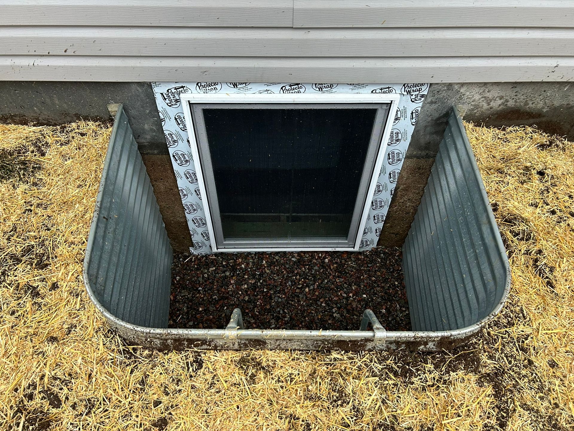 A basement window with a corrugated metal window well installed in the ground against a house foundation.
