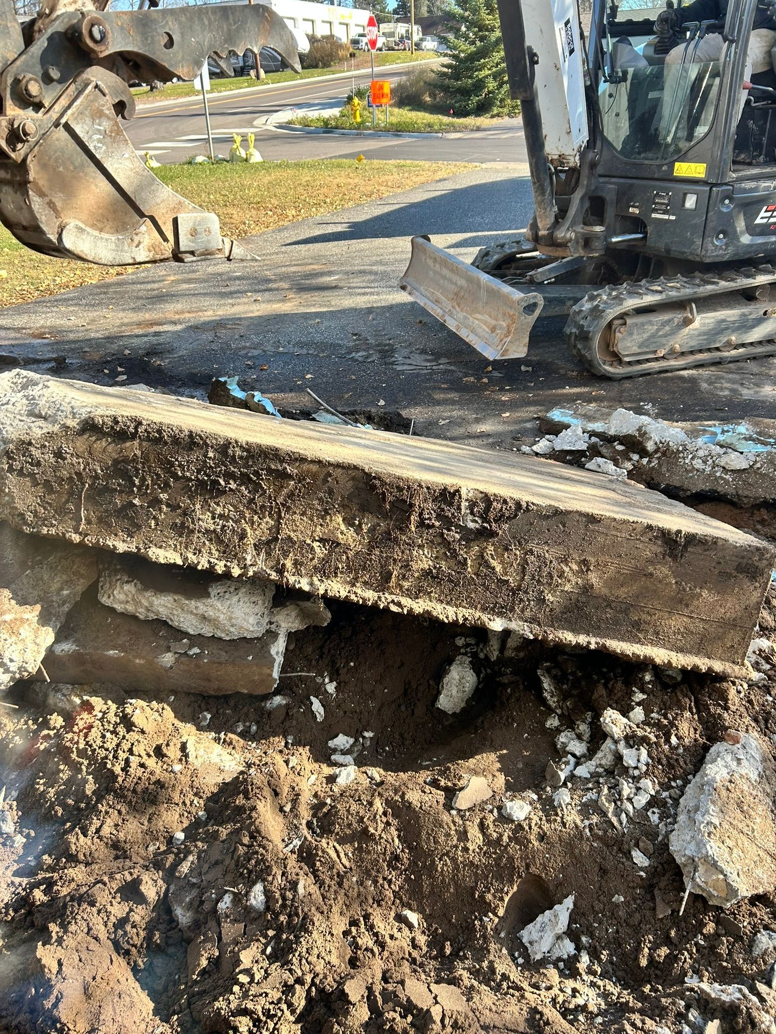 An excavator breaks apart a thick concrete slab at a construction site, exposing the soil underneath.