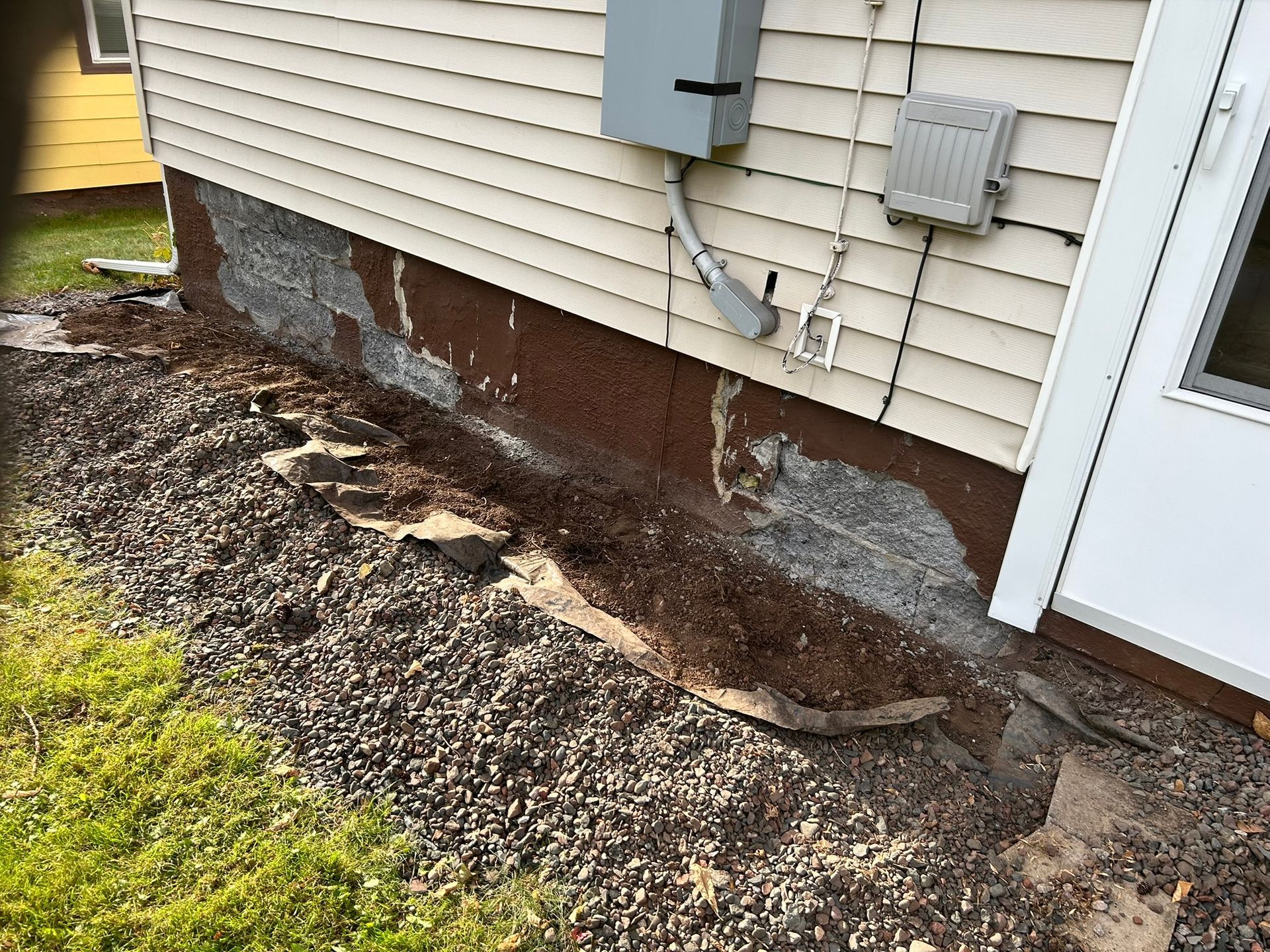 A section of a house exterior showing exposed, crumbling concrete foundation beneath tan vinyl siding.