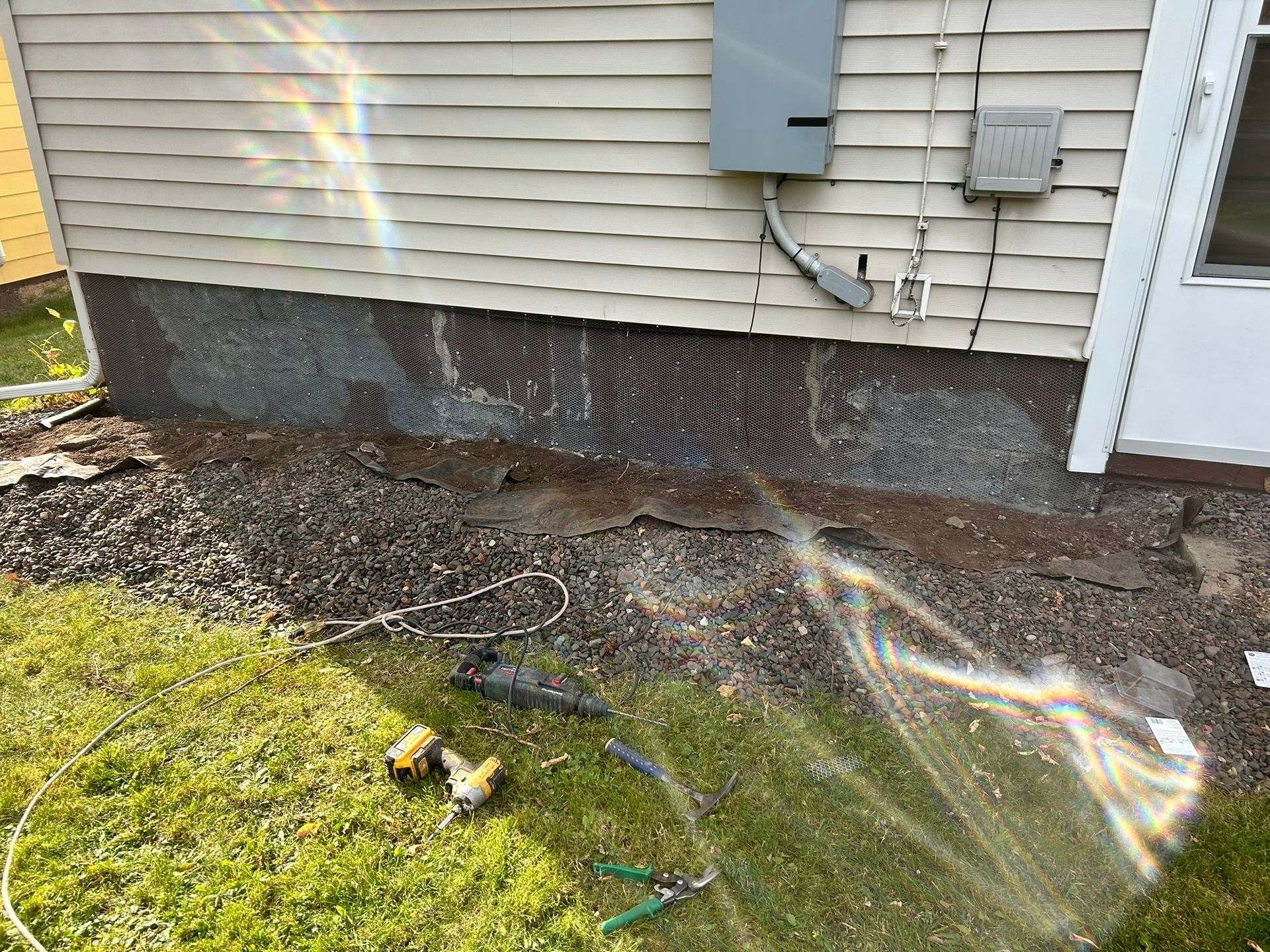A residential exterior showing a concrete foundation with loose mesh, siding, an electrical box, and tools on the grass.