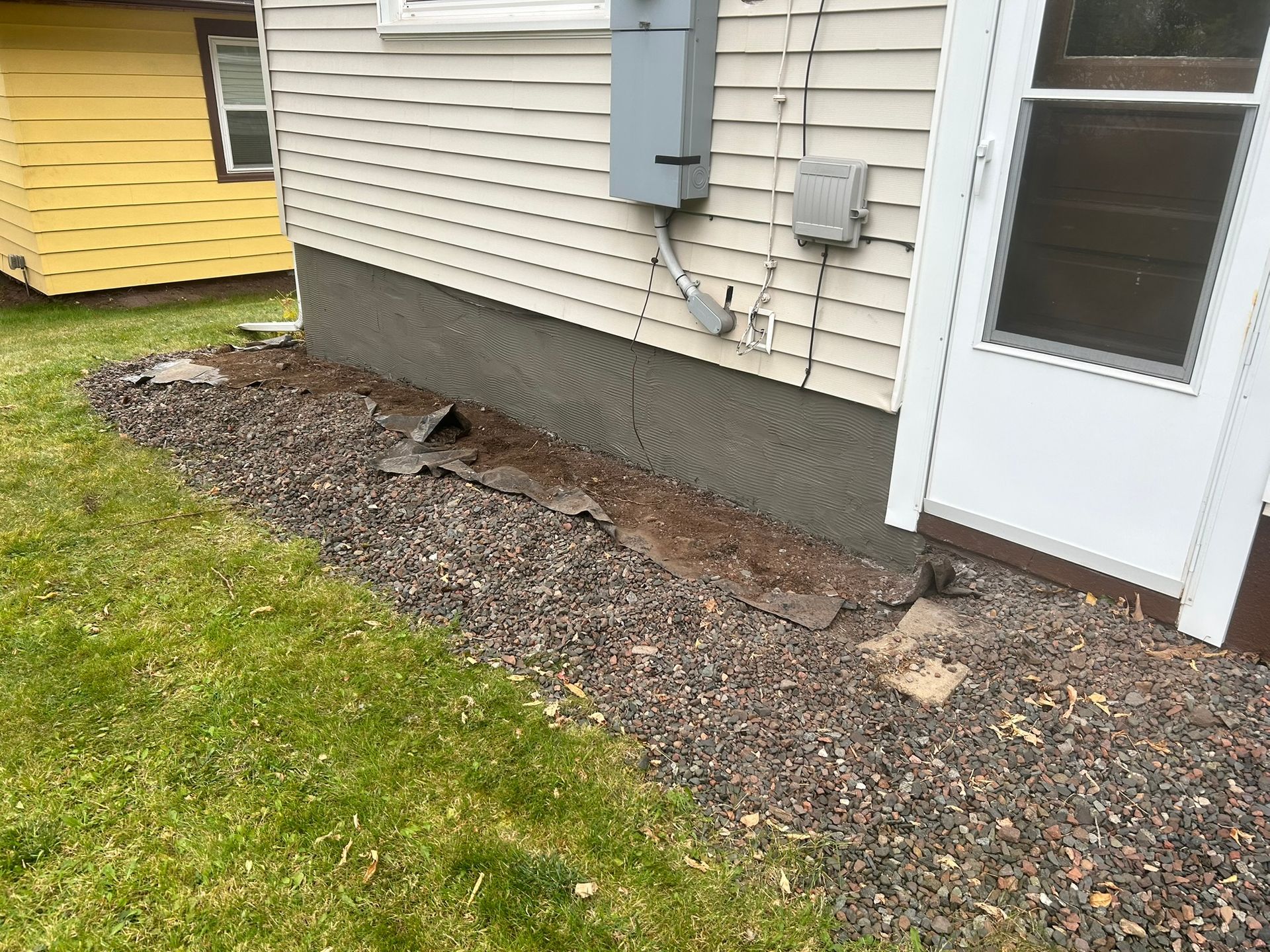 A gravel patch along a house foundation featuring siding, an electrical meter box, and a white door with a storm door.
