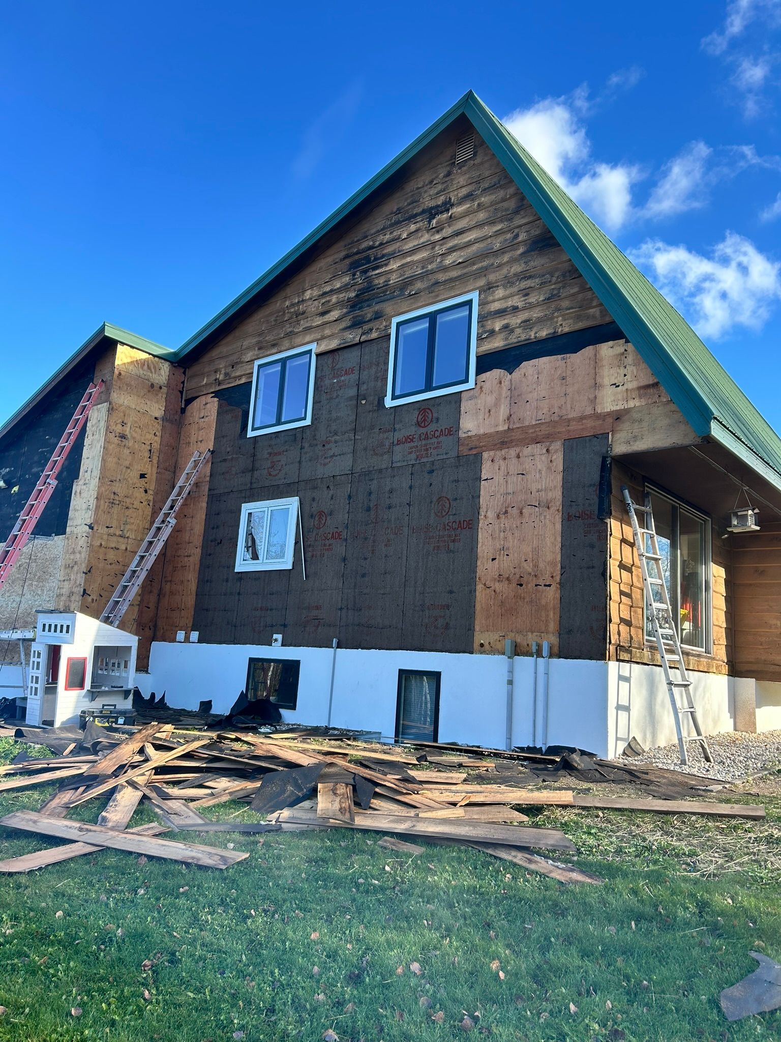 A house under exterior renovation with exposed wood sheathing, black house wrap, and white foundation, seen on a sunny day.