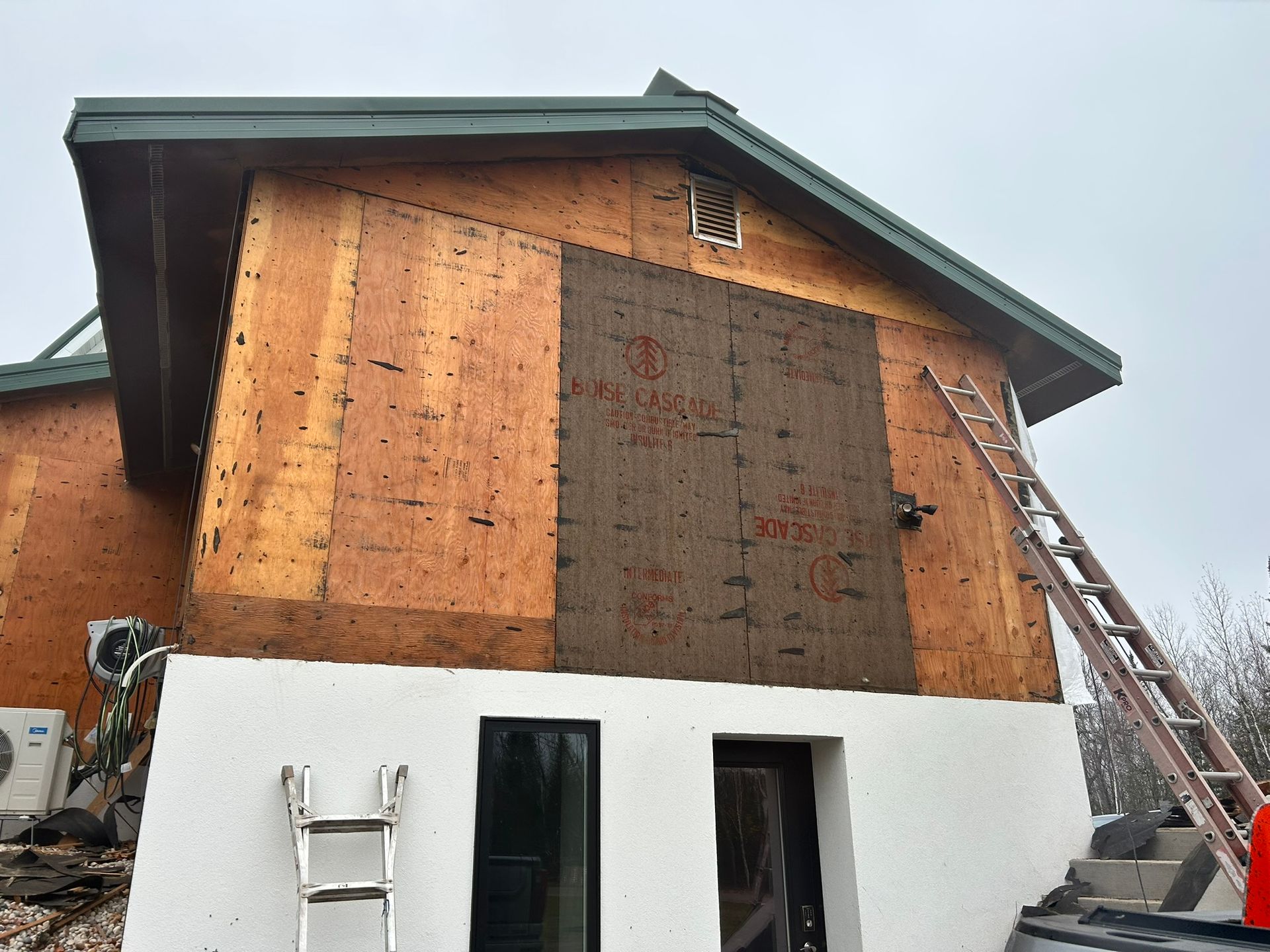 A two-story house under construction with exposed plywood siding, a partially installed dark panel, and a white lower level.