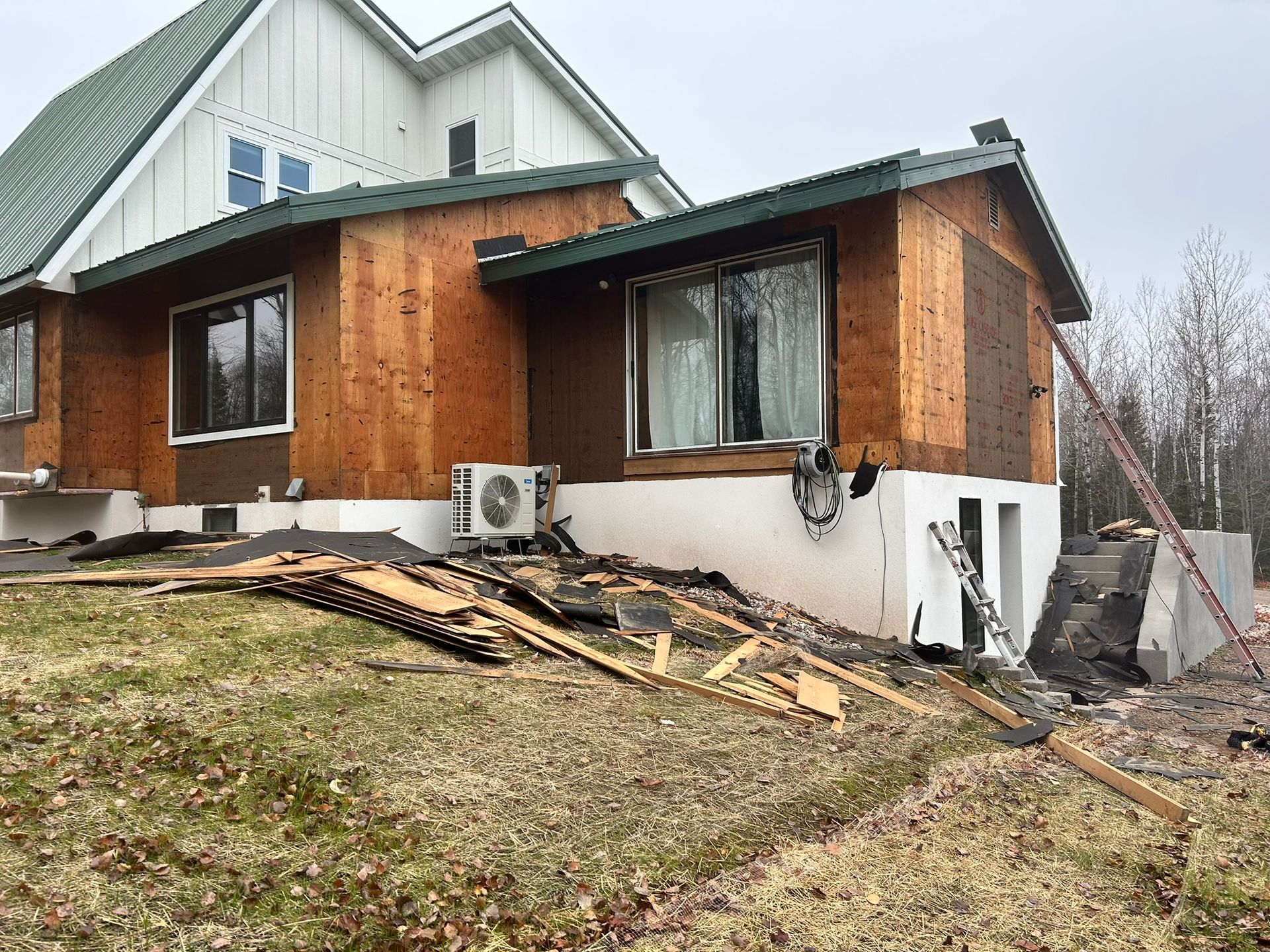A house under exterior renovation with plywood siding, a green metal roof, a white foundation, and scattered lumber.