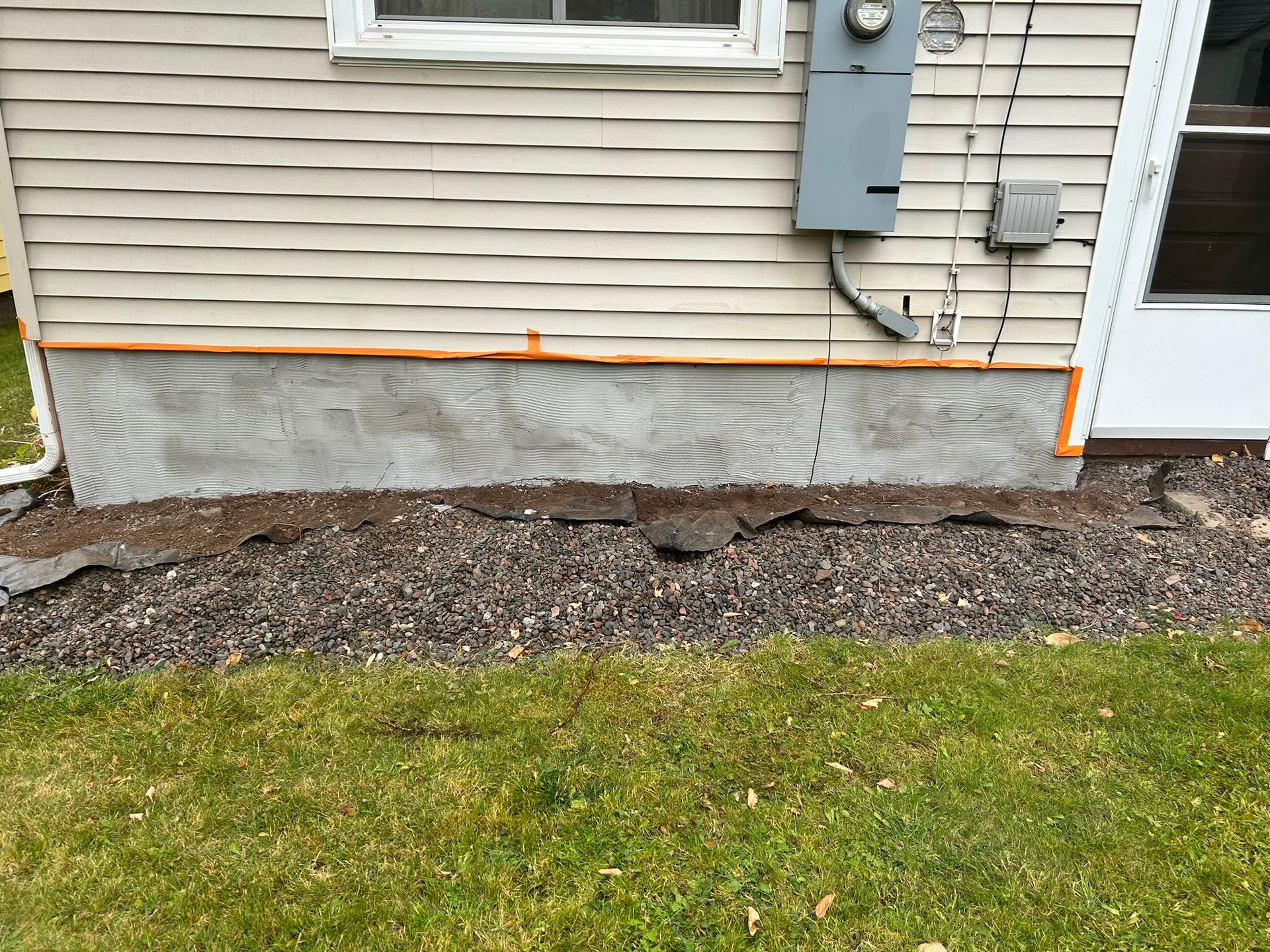 Beige siding above a plywood-covered foundation with orange foam sealant, situated above dark mulch and a grassy lawn.