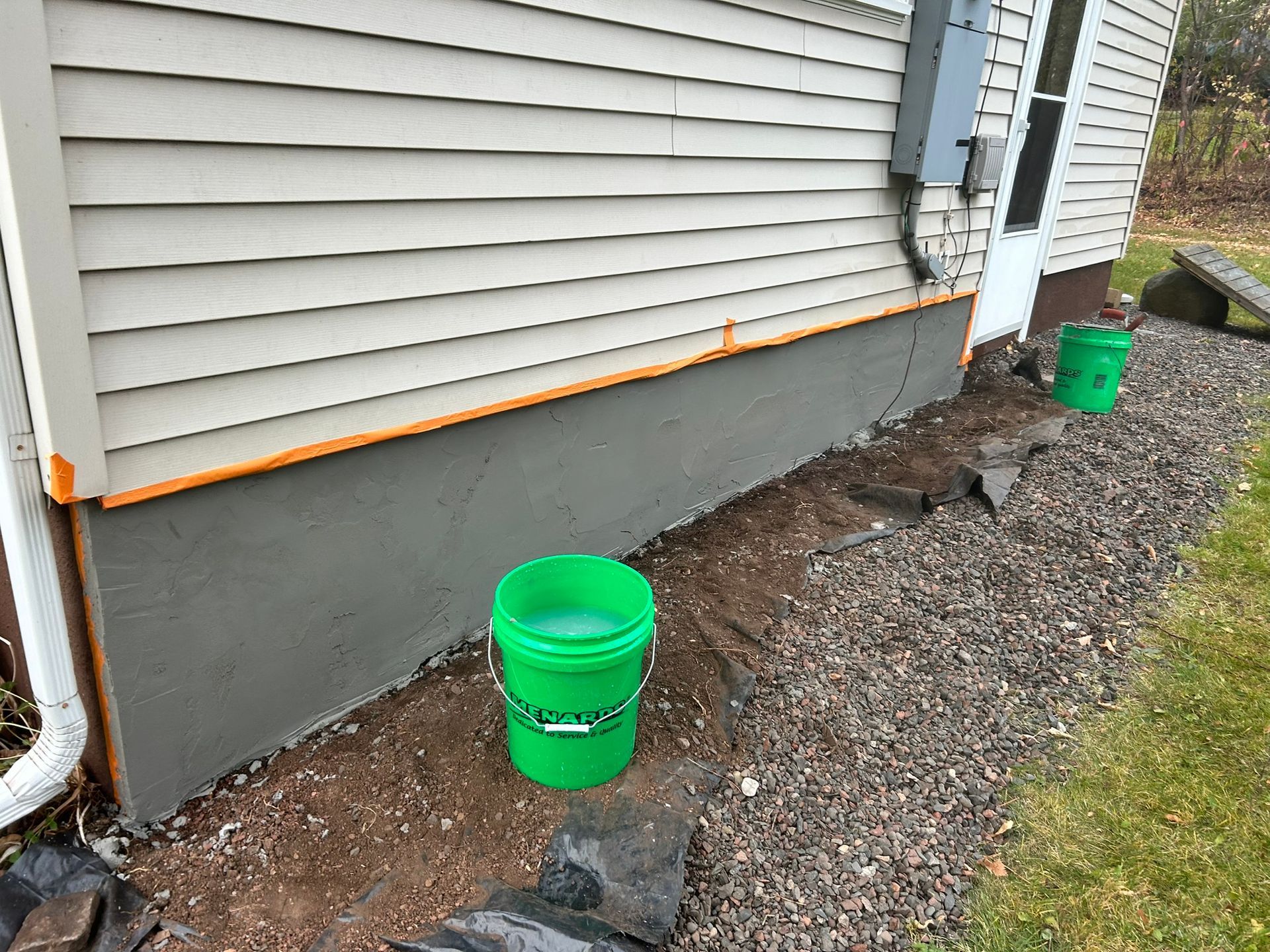 A freshly parged gray foundation wall against a beige-sided house, with green buckets on a gravel ground.