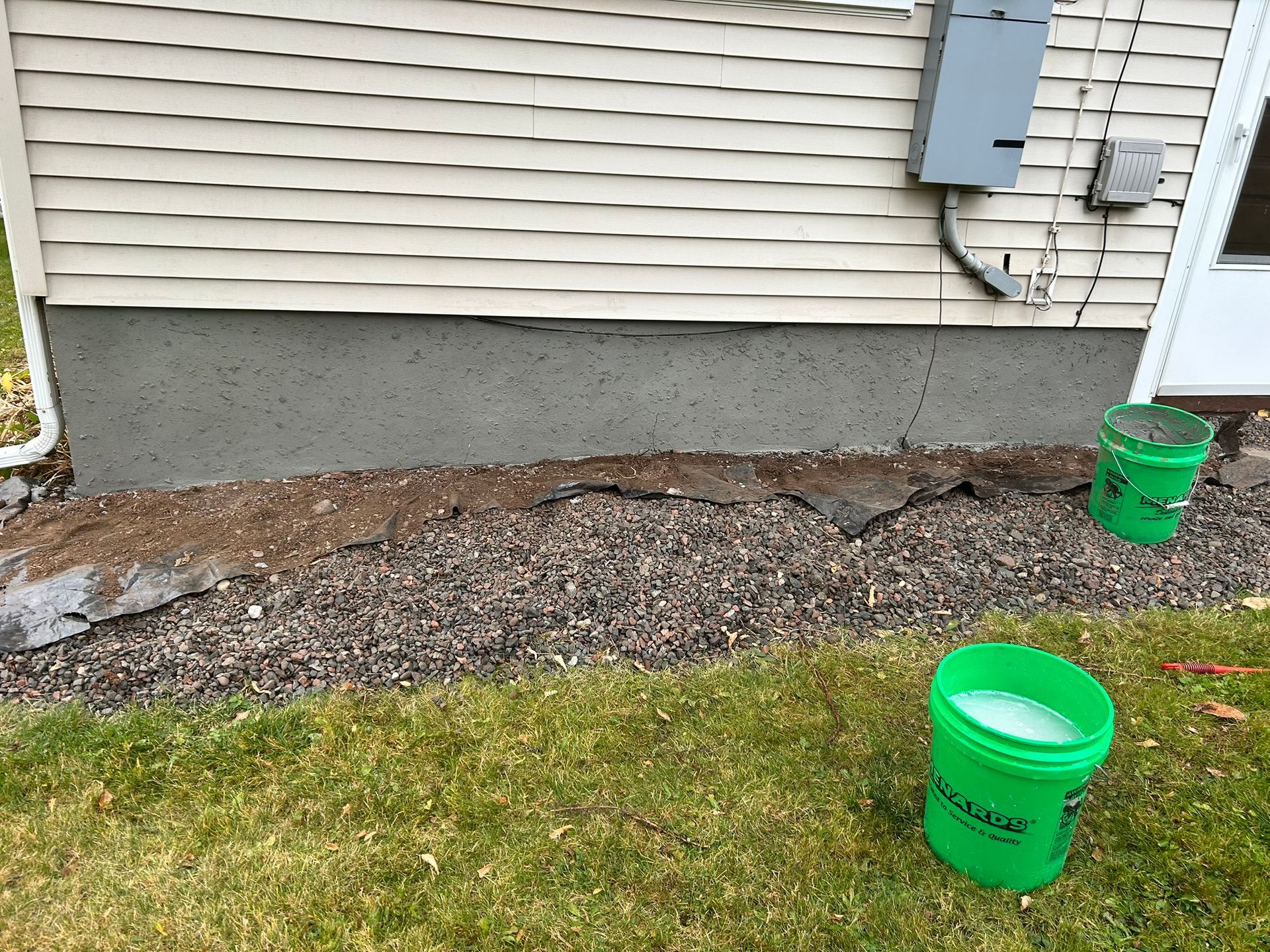 A tan-sided house foundation with a vertical crack, a layer of landscaping fabric, gravel, and two green buckets outside.