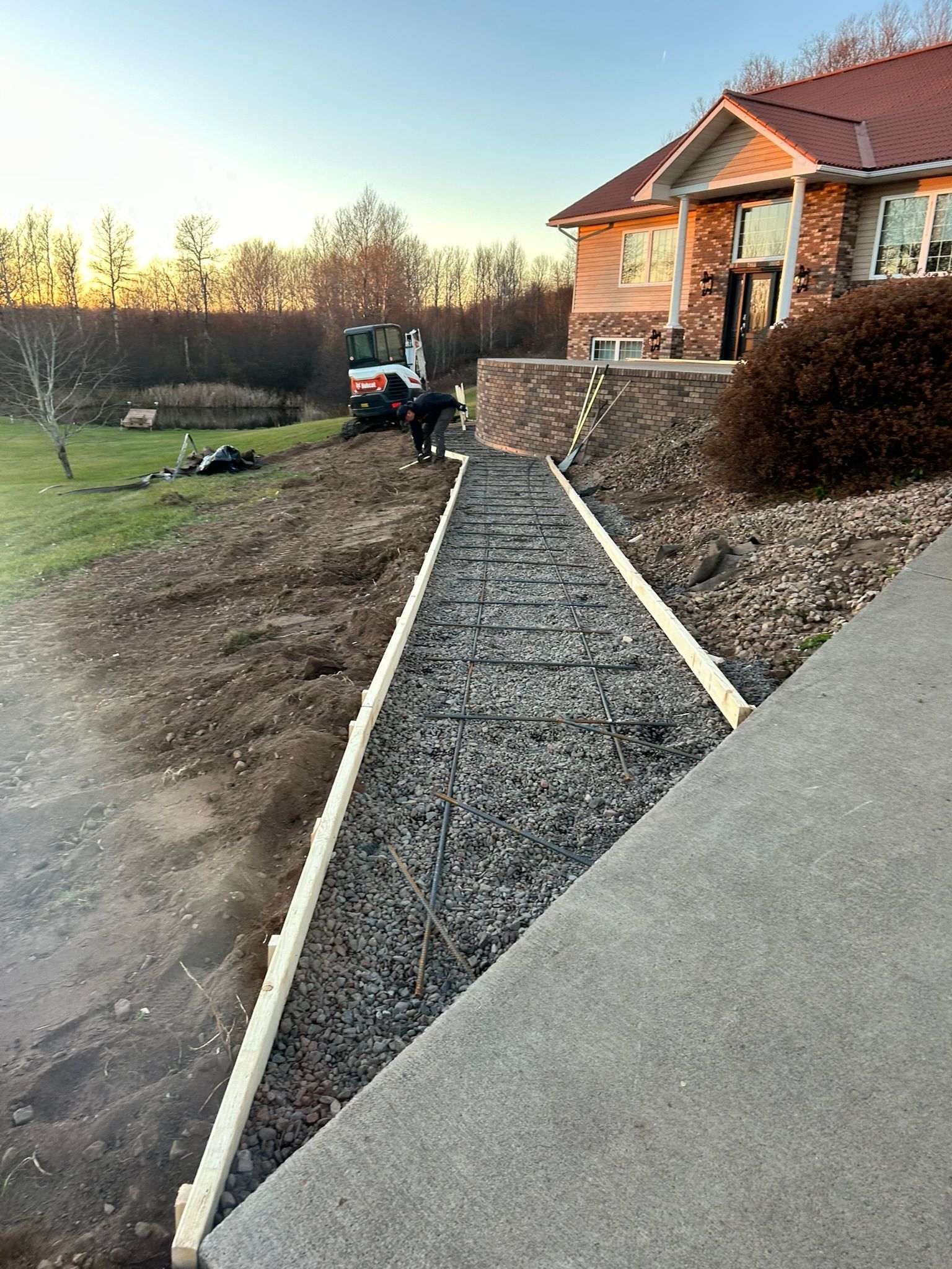A stone path under construction with wooden forms leads toward a house with a brick exterior at sunset.