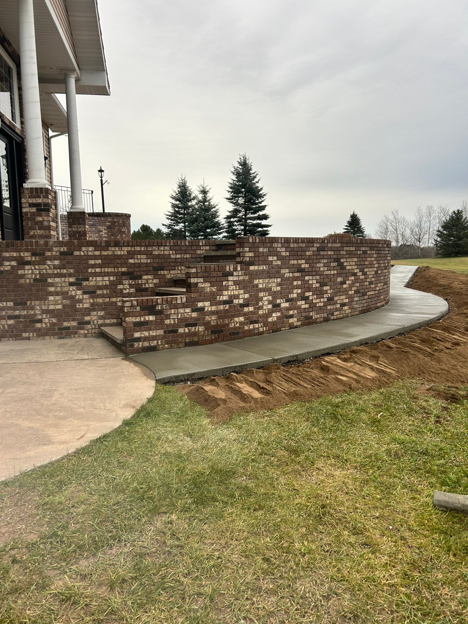 Curved brick retaining wall with stone steps leading from a patio to a concrete path on a grassy lawn.