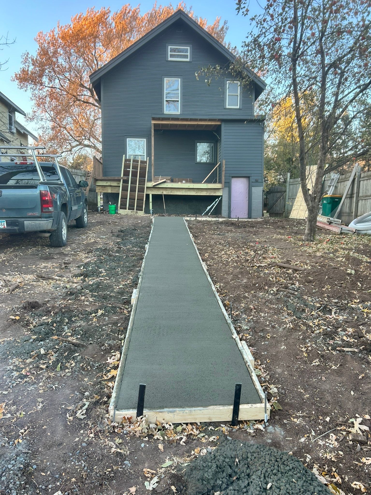 A fresh strip of wet concrete sidewalk leads toward a dark-sided two-story house with a wooden deck under construction.