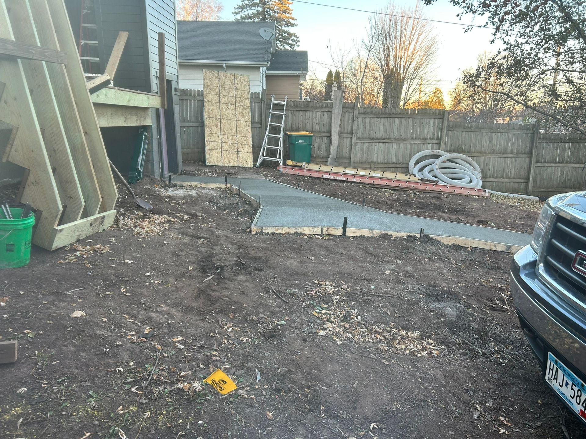 A newly poured concrete walkway in a backyard, near a construction area with a ladder and a wooden fence.