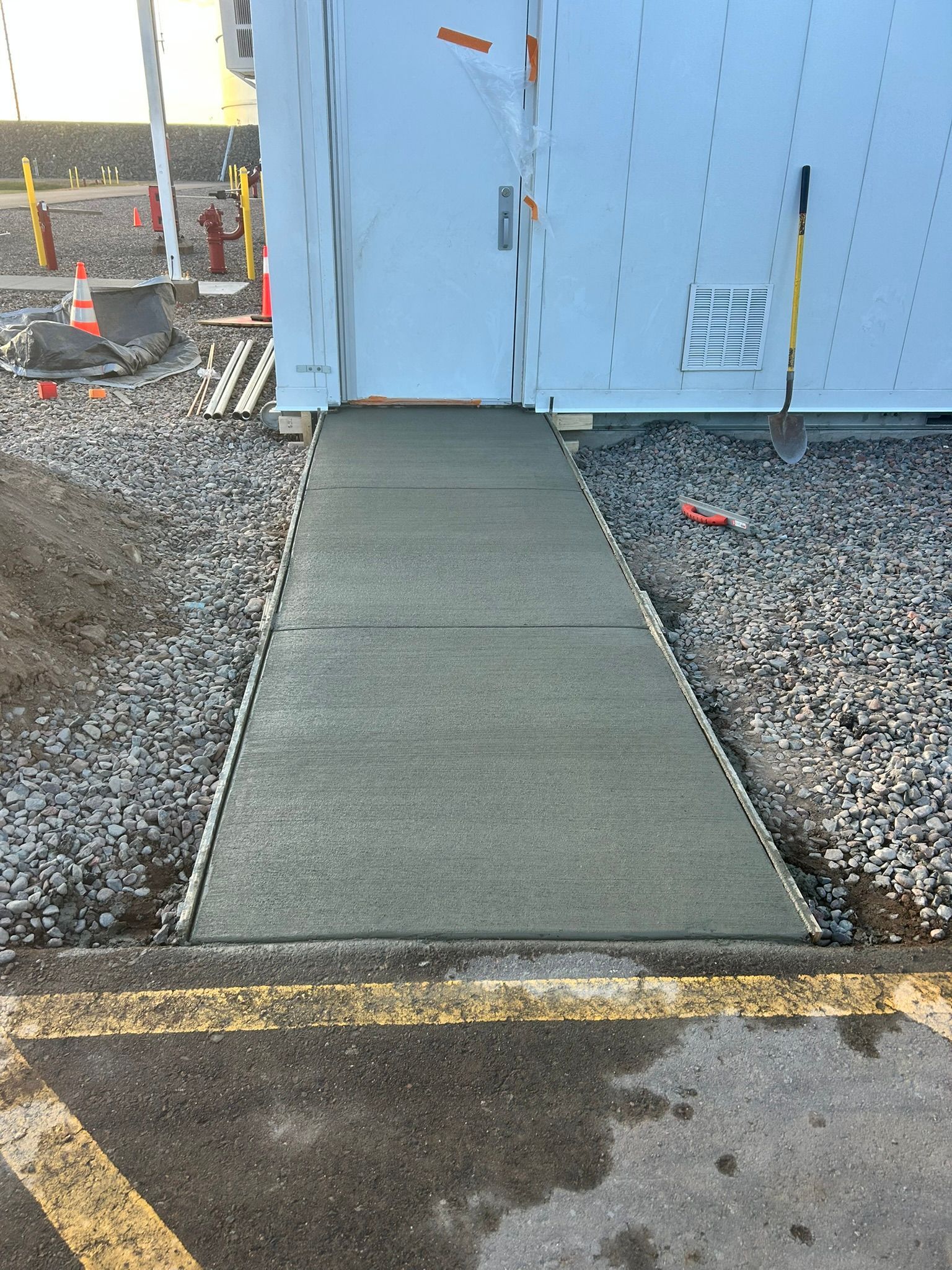 A concrete walkway leading to a doorway on a white building exterior, flanked by gravel and construction equipment.