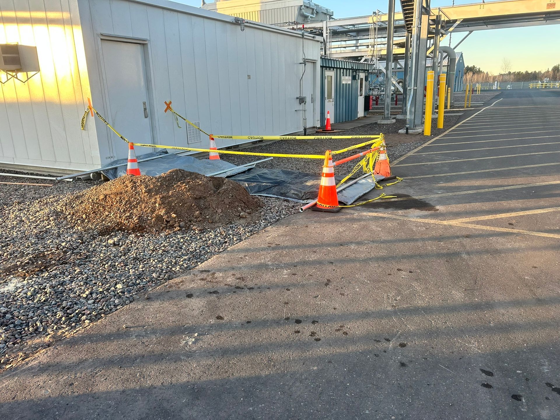 A pile of gravel sits outside a white building, cordoned off by traffic cones and yellow caution tape on an asphalt lot.