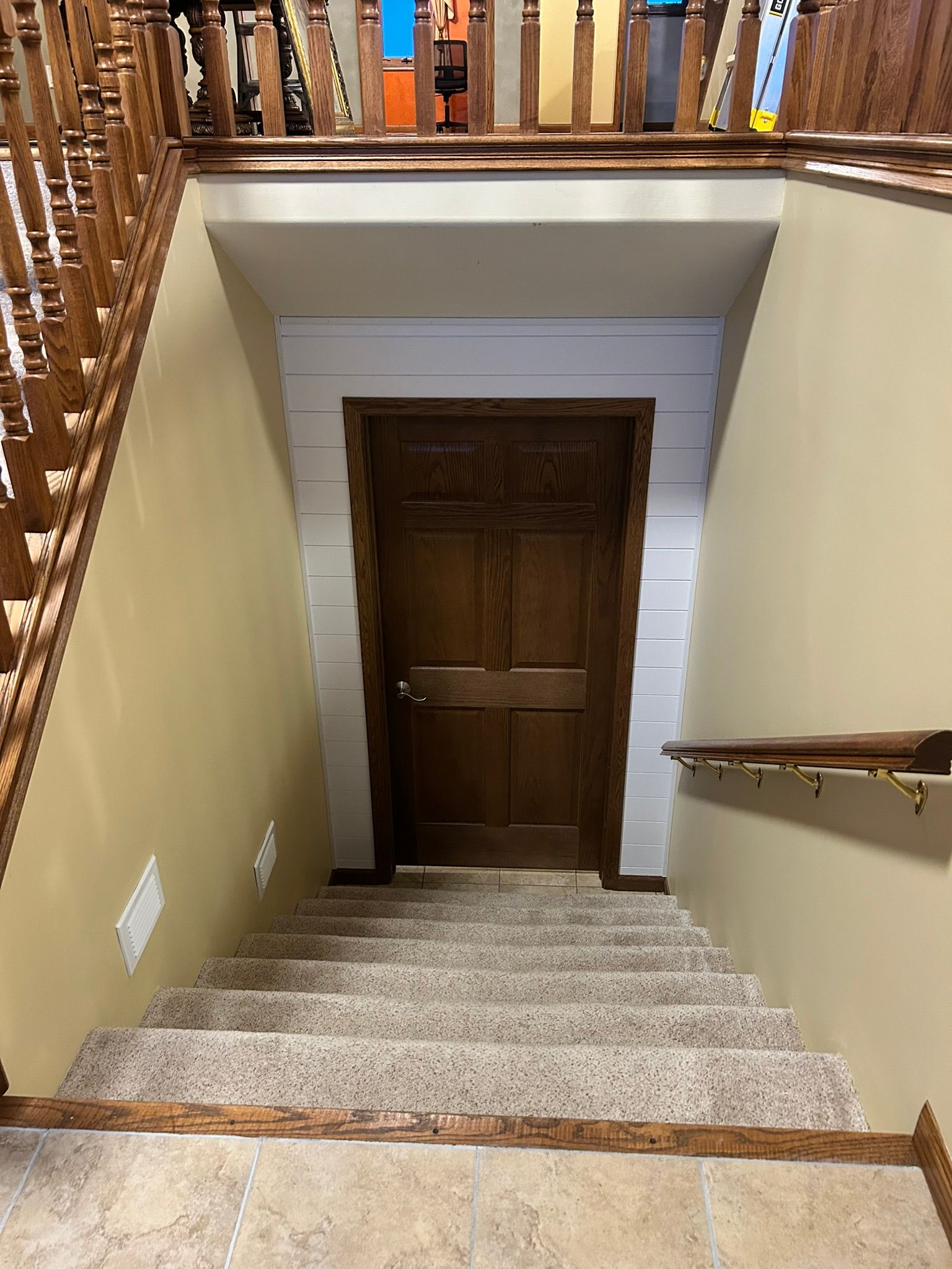 A staircase with beige carpet descends toward a dark wood door framed by white shiplap walls.