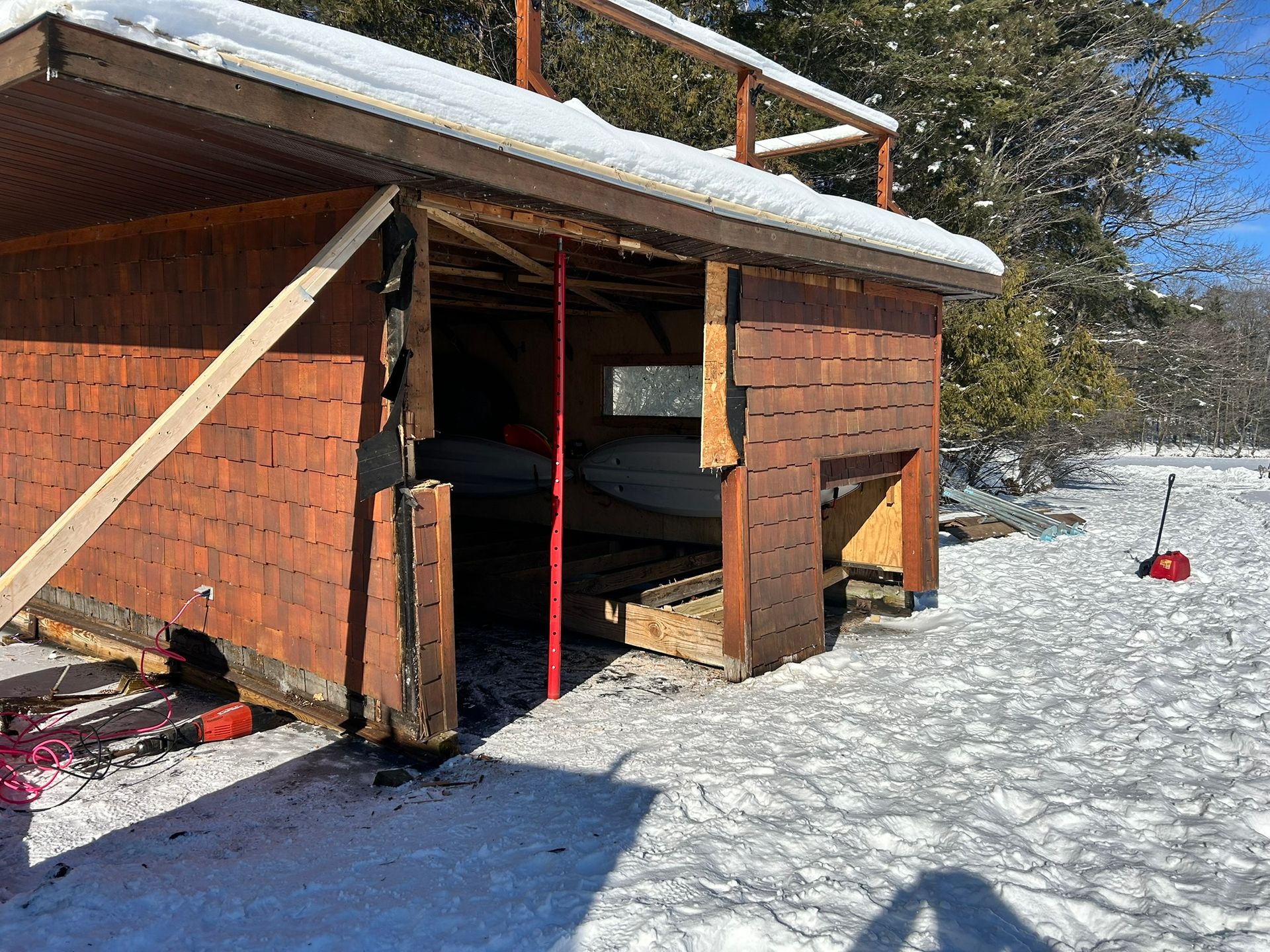 A brown wooden structure with an open side and a snow-covered roof, braced by a temporary support, stands on a snowy field.
