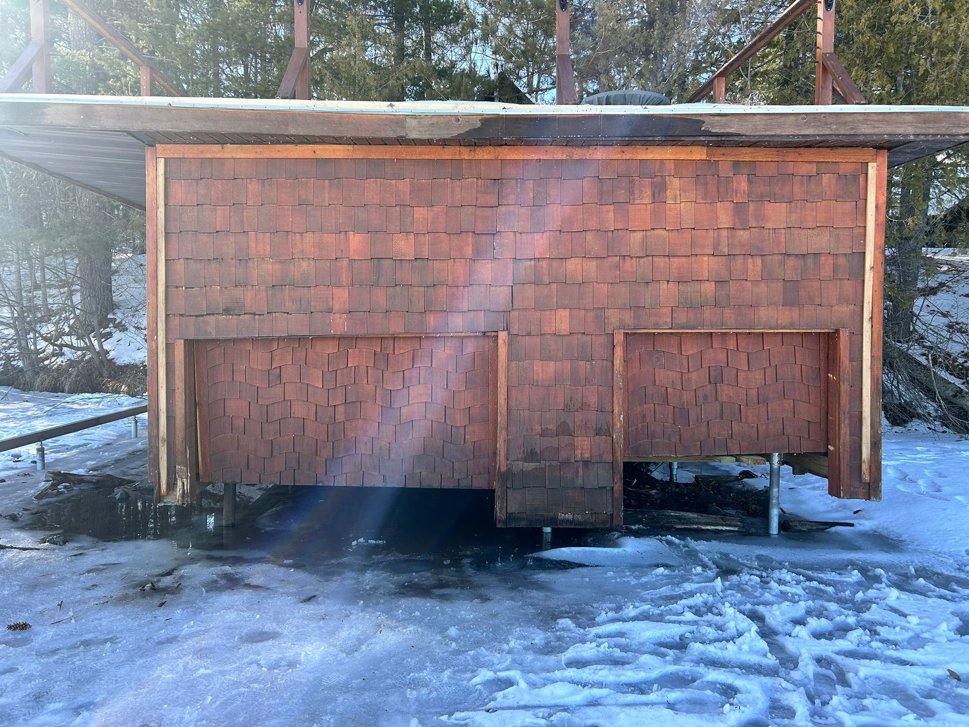 A wooden cabin facade with cedar shake shingles, elevated on metal posts above frozen, snow-covered ground.
