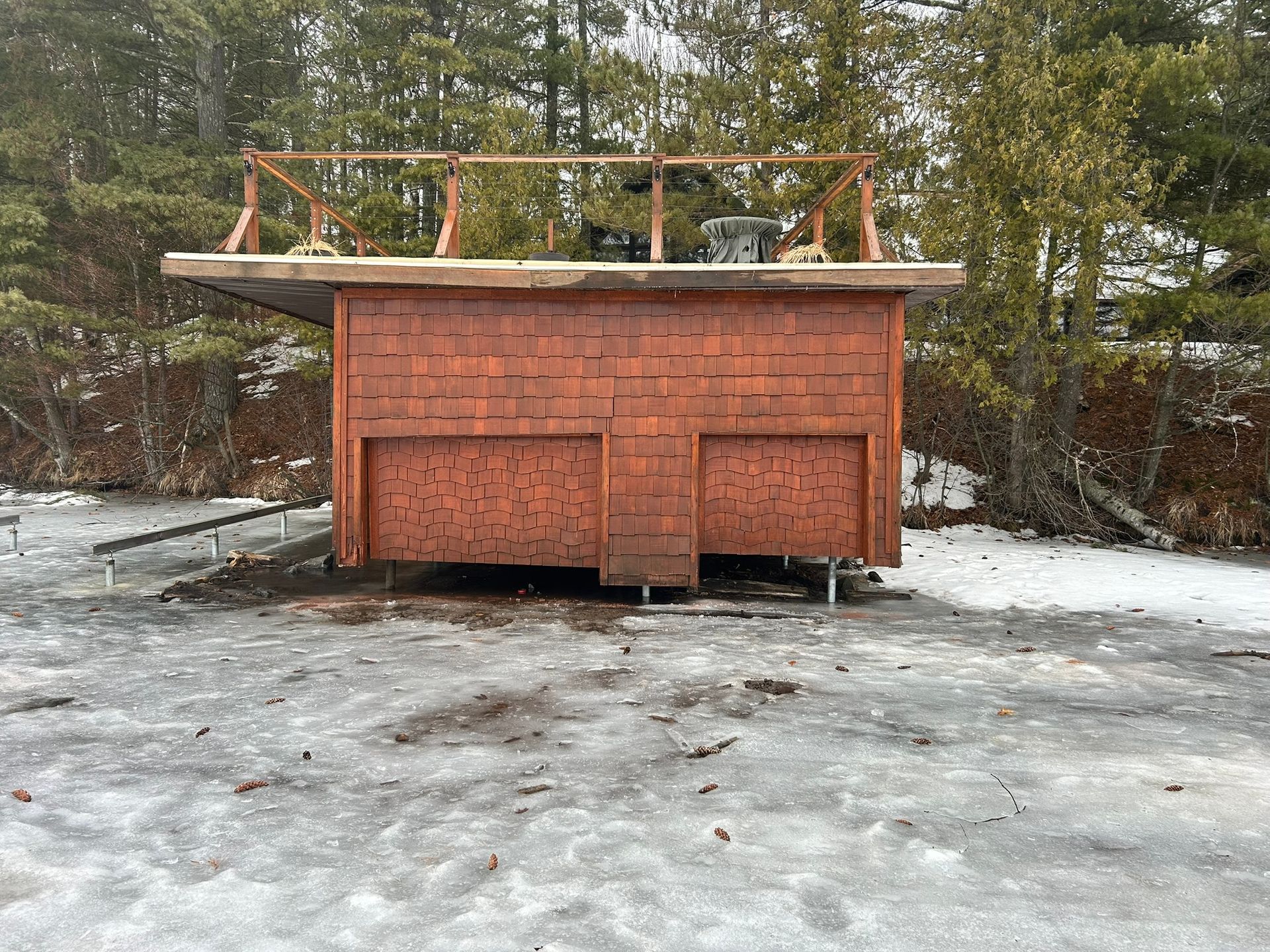 A wooden structure with a flat, unfinished roof frame sits on a frozen, snow-covered surface near a forest.