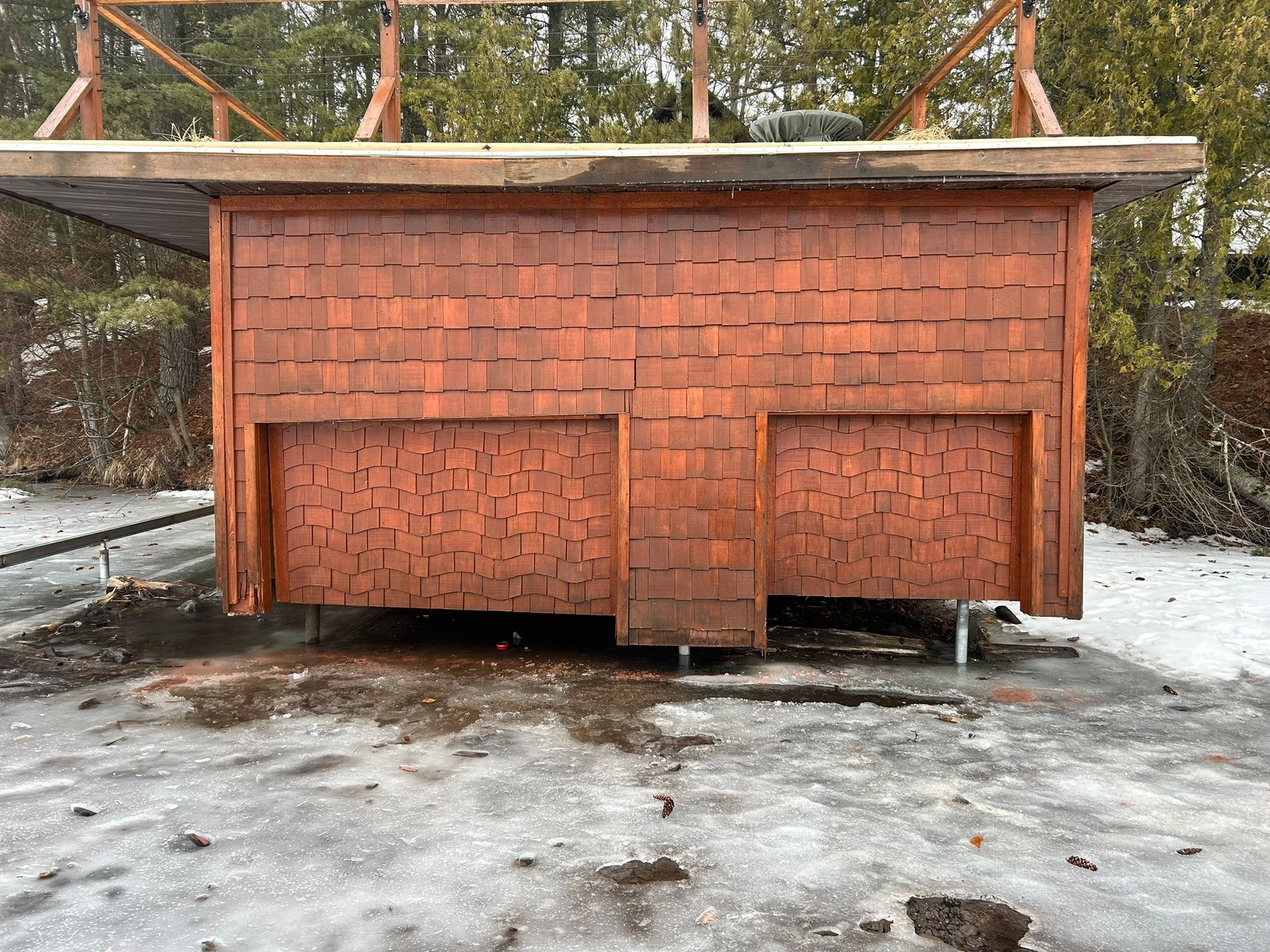 A small, rectangular structure with reddish-brown wood shingle siding stands on posts over an icy, snow-covered surface.