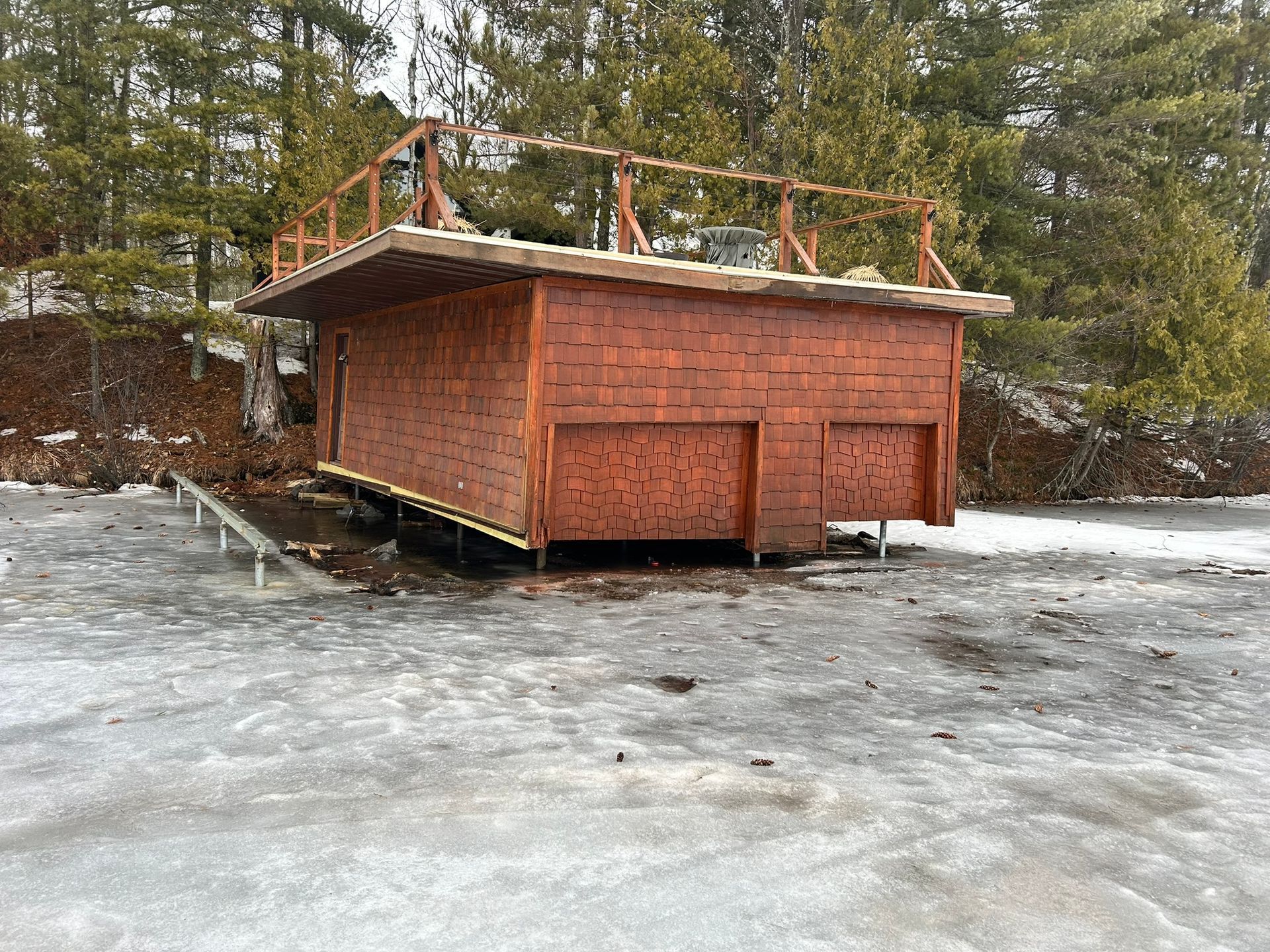 A brown, rectangular shed-like structure with a rooftop deck stands on a frozen, icy surface near a treeline.