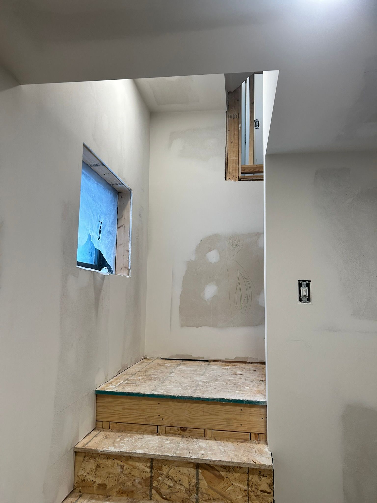 An unfinished stairwell with bare plywood steps and drywall, leading up to a landing with a small window.