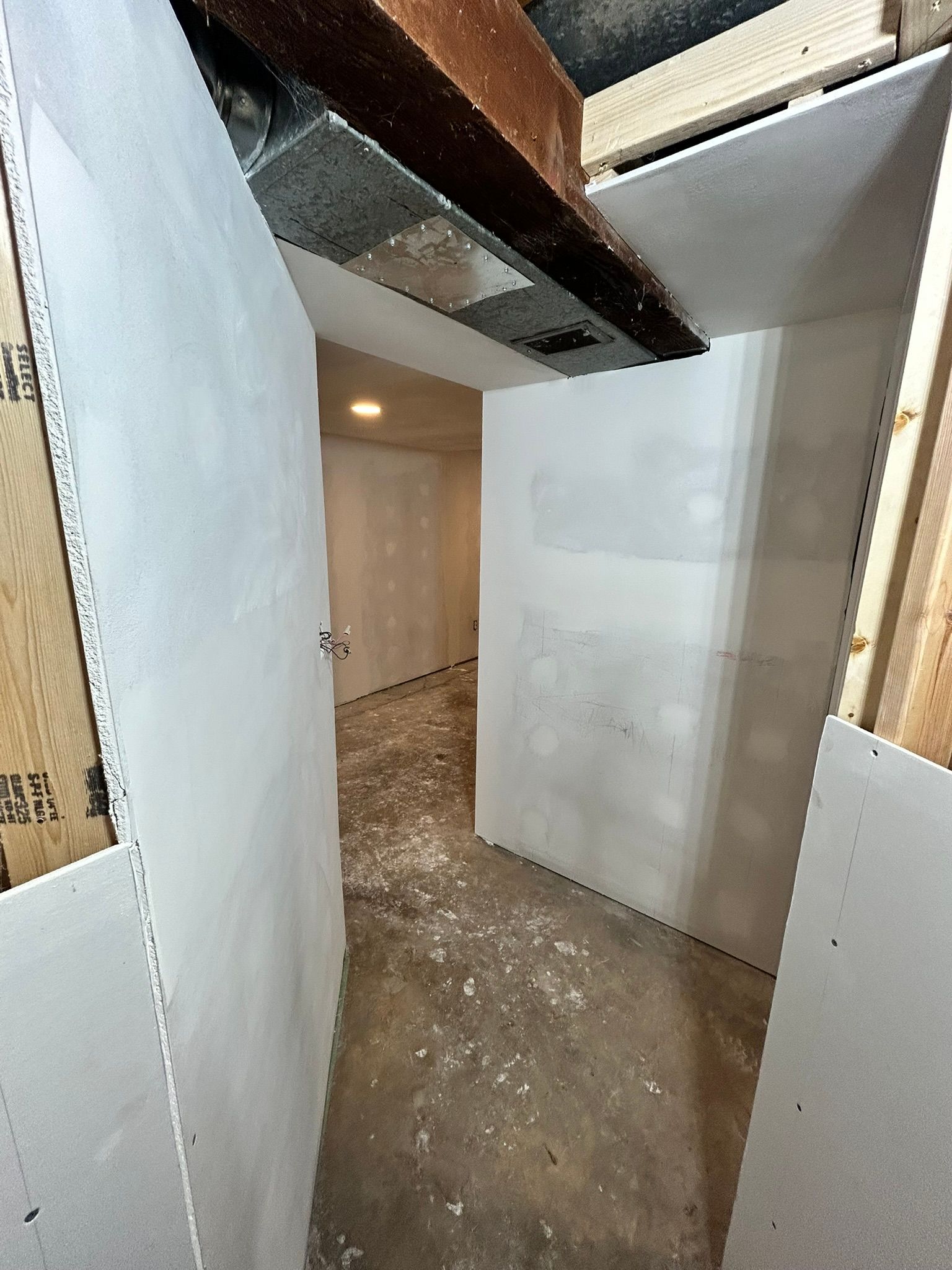 A perspective view of a narrow, unfinished basement hallway with newly installed white drywall, showing a concrete floor.
