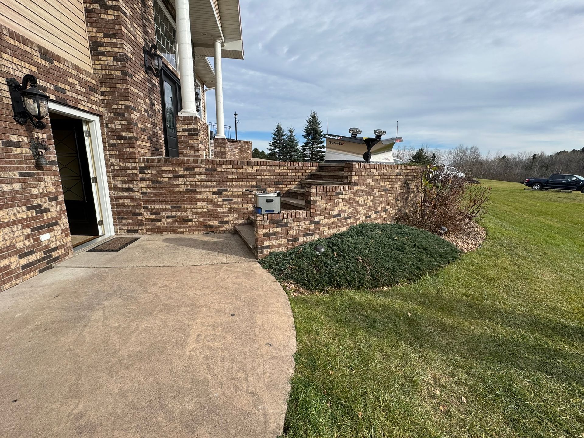 A brick house exterior with a patio, steps leading to a raised porch, and a grassy lawn under a cloudy blue sky.