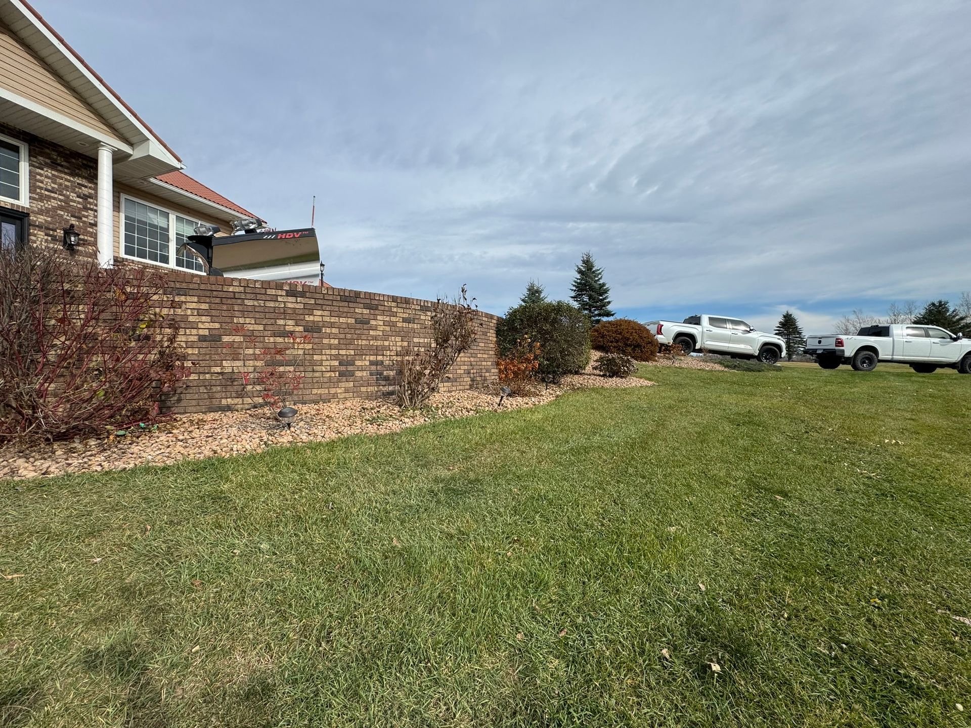 A brick house with a terraced retaining wall overlooks a grassy hill with two white pickup trucks parked in the distance.