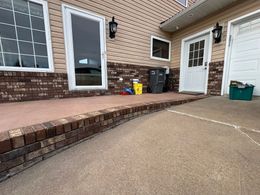 A residential exterior with a brick-trimmed concrete patio, white door and window frames, and beige siding.