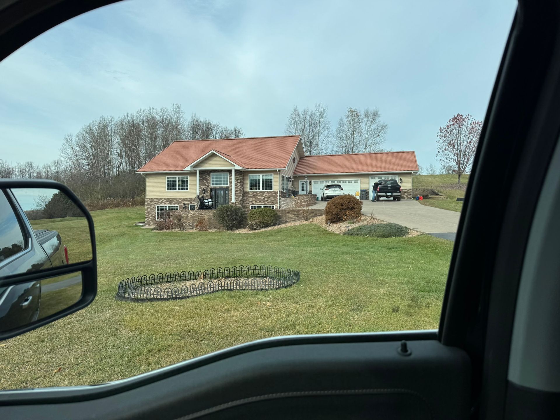 A house with a reddish-brown roof, stone foundation, and a sprawling green lawn, viewed from inside a vehicle.