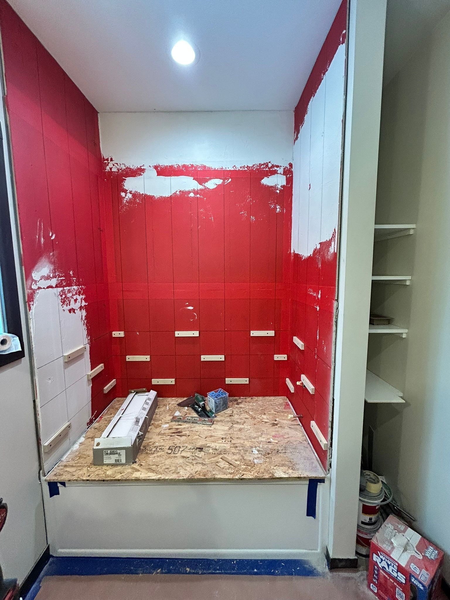 A shower niche under construction with red waterproof membrane on the walls, a wooden base, and adjacent built-in shelves.