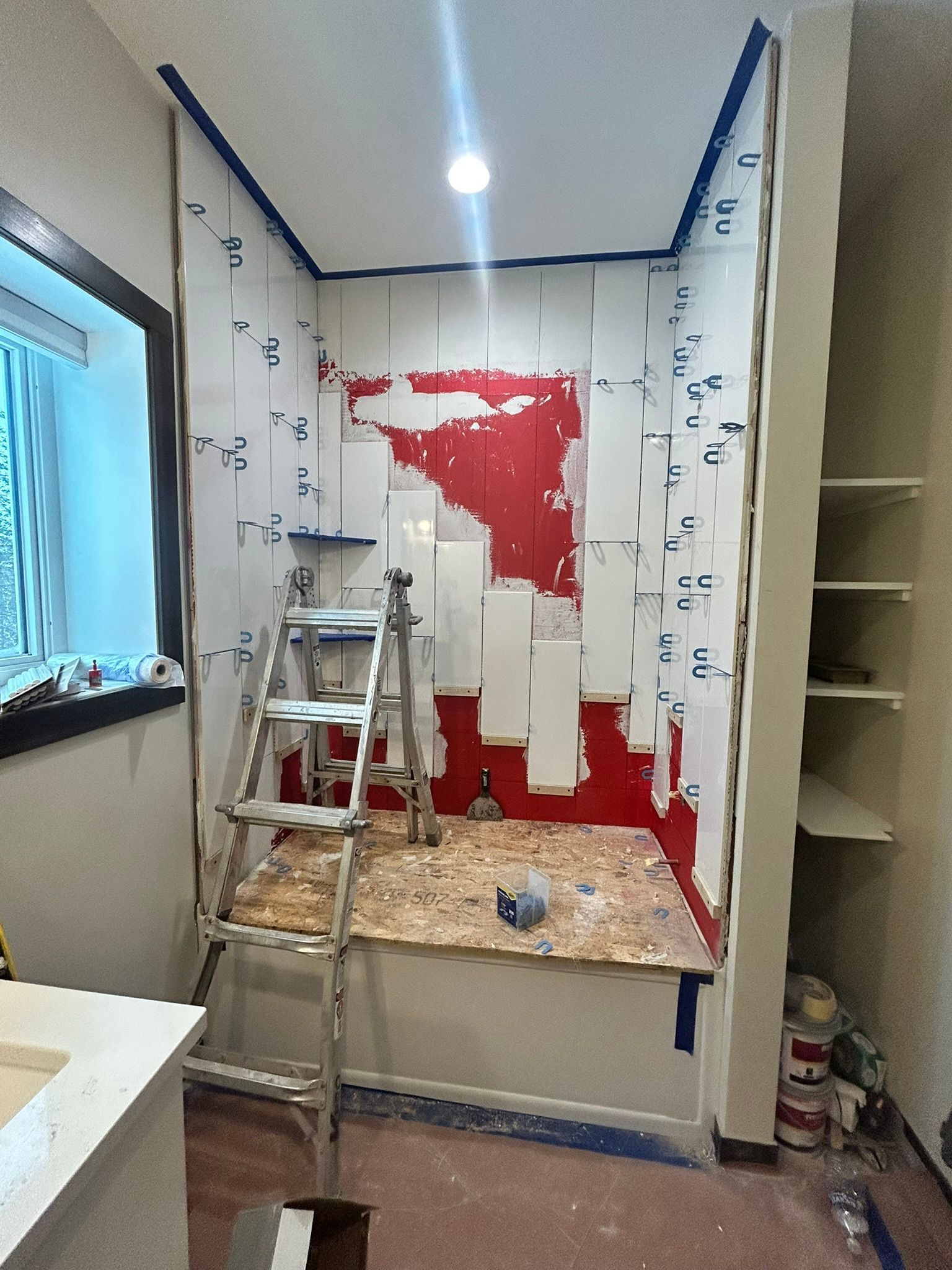 A step ladder stands in a bathroom alcove where white shower wall panels are being installed over a red waterproofed area.