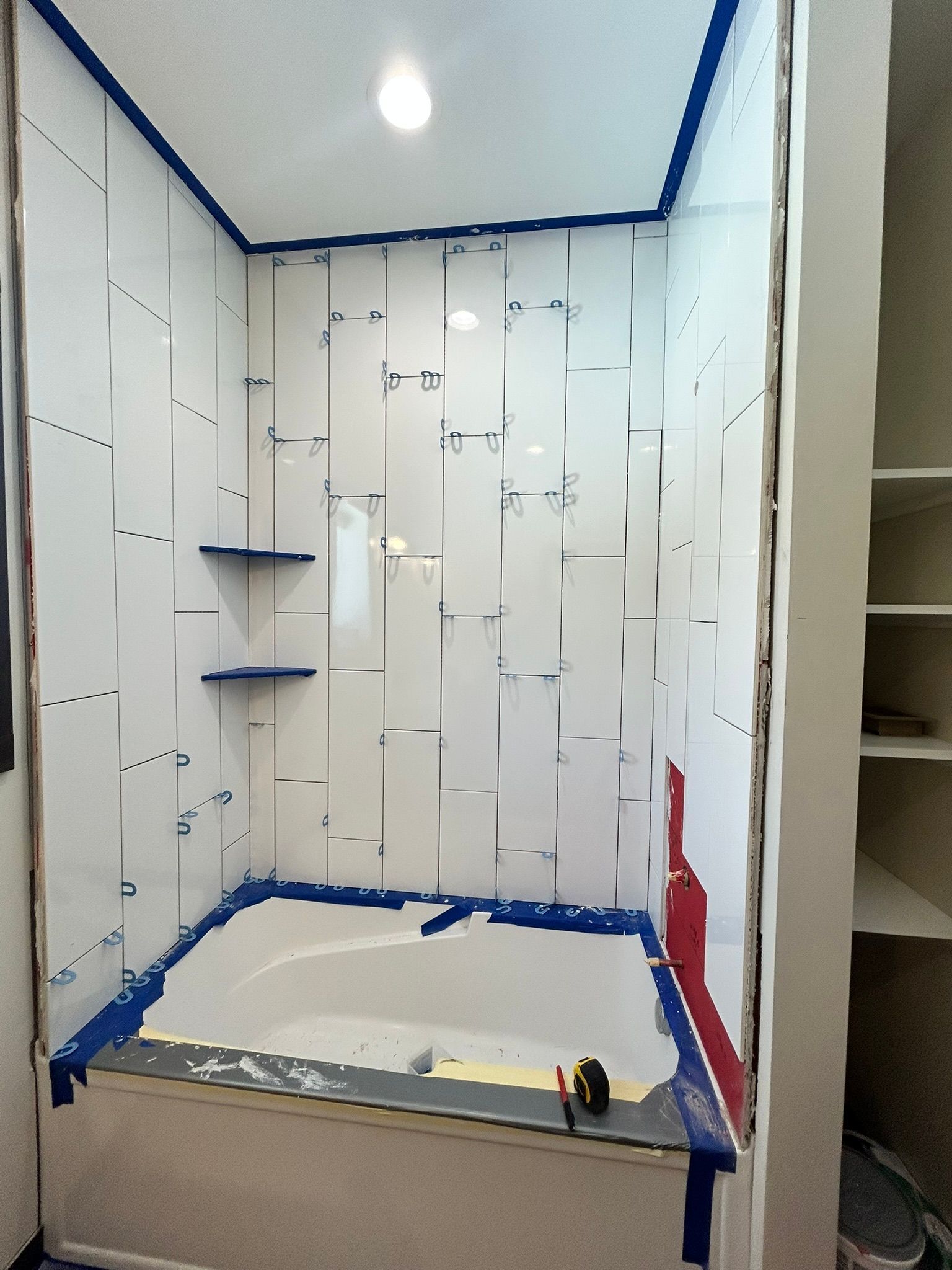A white shower surround being installed in a tiled alcove, featuring blue painter's tape and two built-in corner shelves.