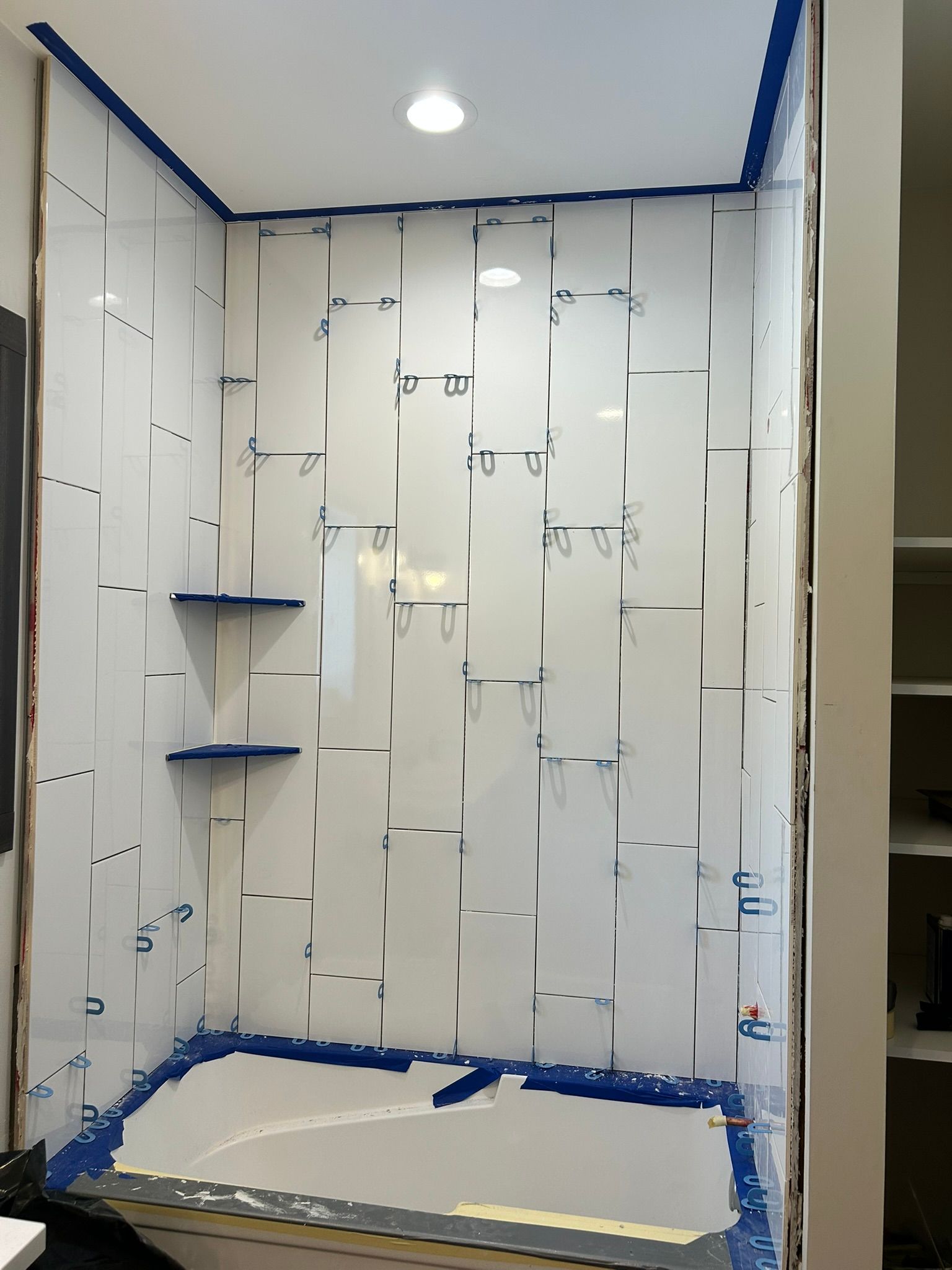 A white shower surround with chrome edge trim, tape, and two installed corner shelves during a bathroom renovation.