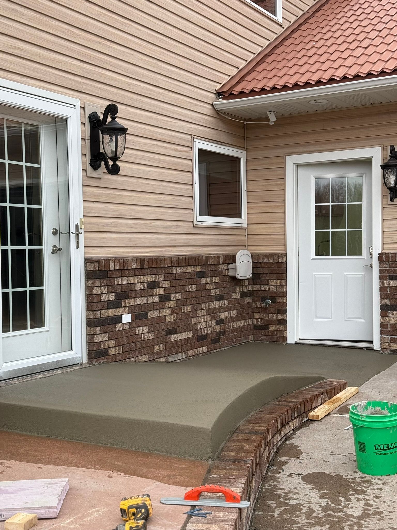 Newly poured concrete porch in front of a house with tan siding and a brick base, beside a green bucket and tools.