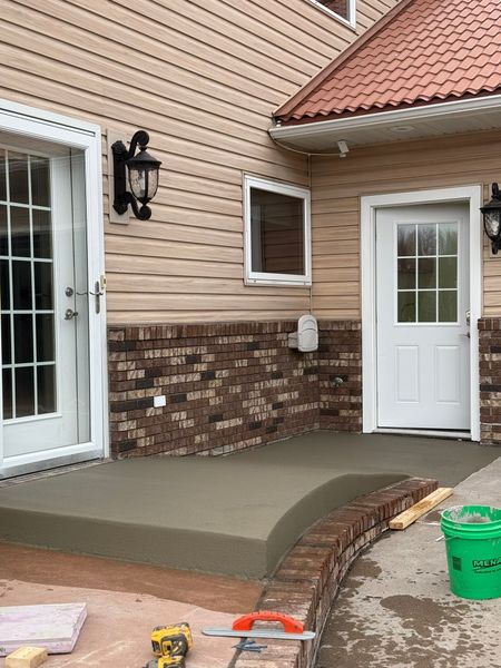 Newly poured concrete porch in front of a house with tan siding and a brick base, beside a green bucket and tools.