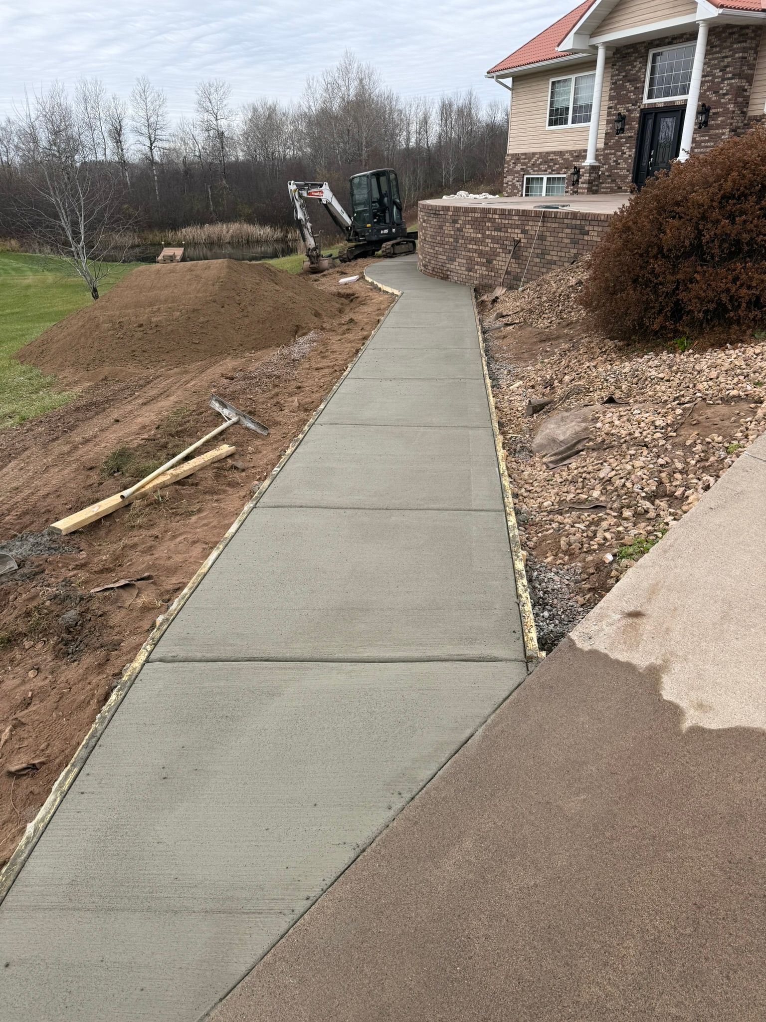 A newly poured concrete walkway leads toward a house entrance with an excavator parked in the background.