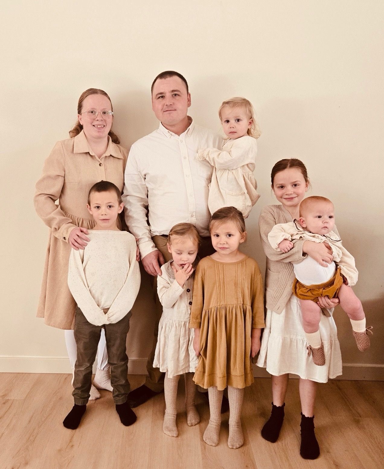 A family of eight posing together against a plain wall indoors, dressed in coordinating beige and earth-toned outfits.